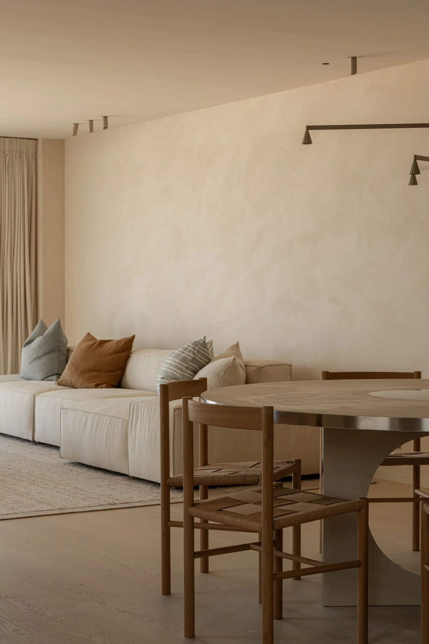 Minimalist living room with a cream sectional sofa complemented by pastel-colored throw pillows and a wooden dining table with matching chairs.