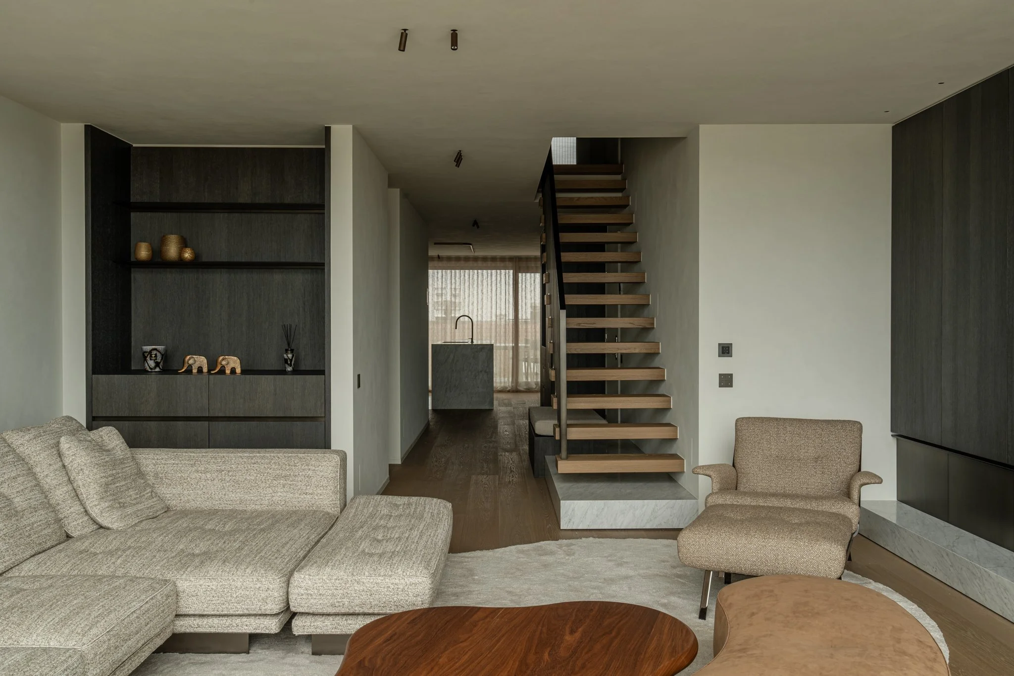 Living room with beige sectional sofa, armchair, wooden coffee table, open staircase, and dark cabinetry, with a kitchen in the background.