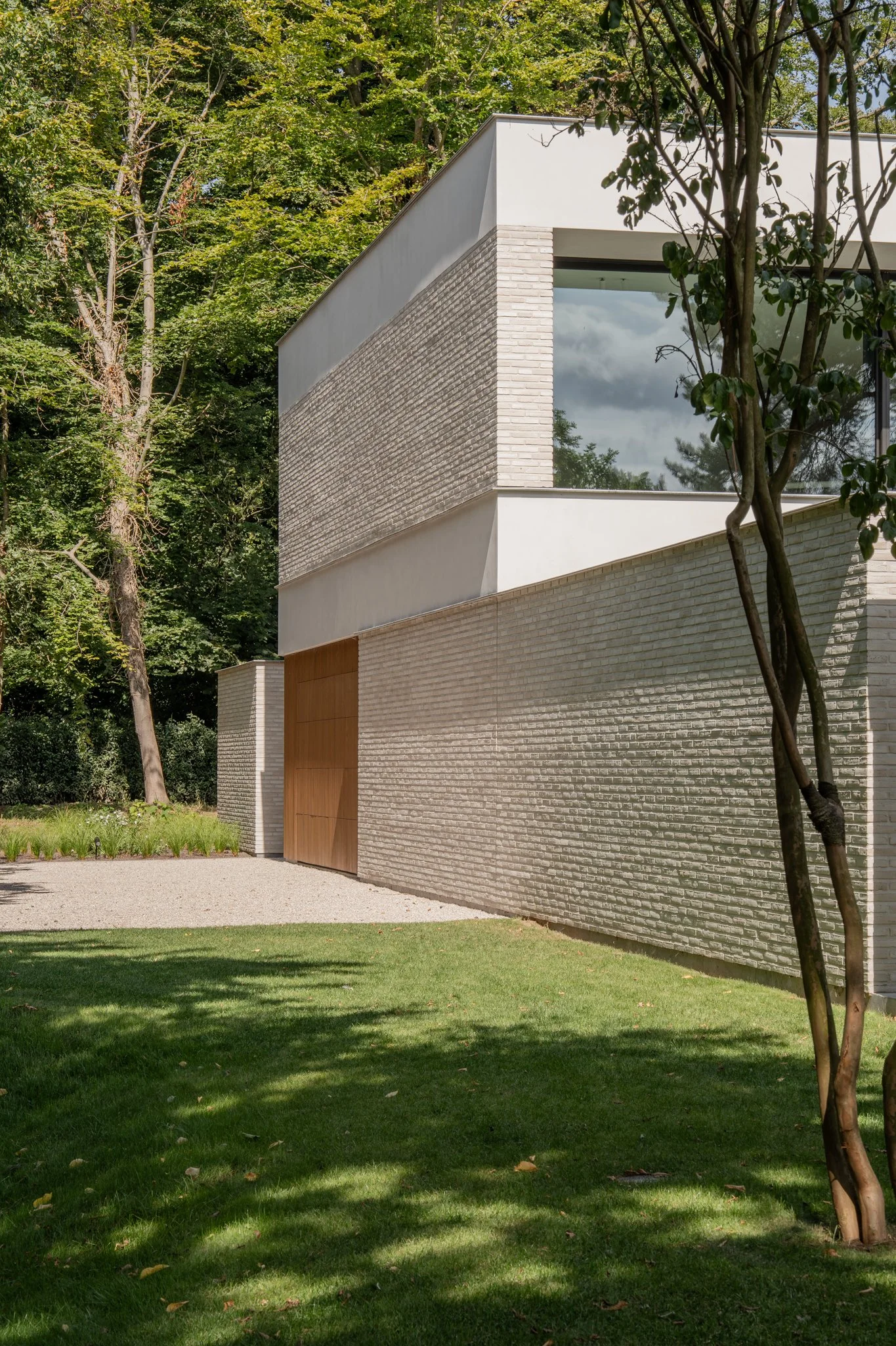 Modern house with gray brick and white walls, wooden door, surrounded by trees and grassy yard.