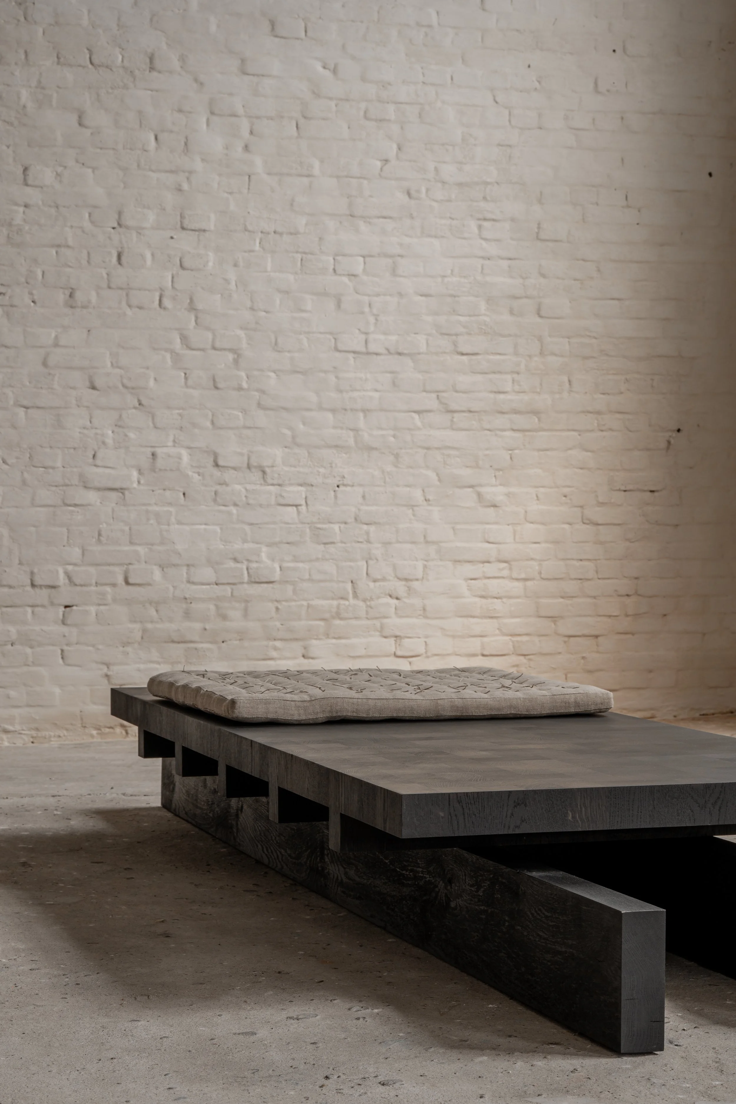 A minimalist low wooden bed with a gray cushion on top, set against a white brick wall in a simple, industrial-style room.
