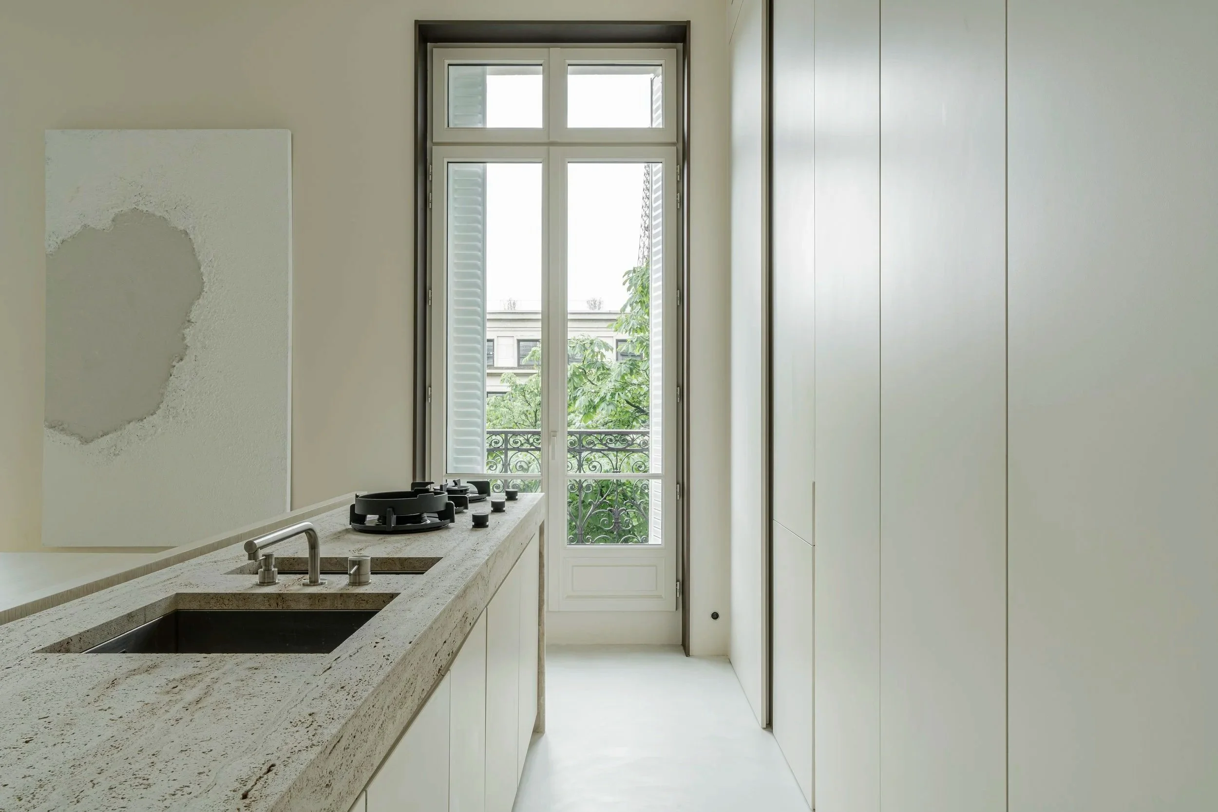 A minimalist kitchen with a speckled white countertop, a built-in sink, and two stovetop burners. There is a large window or door with a balcony and some greenery outside.