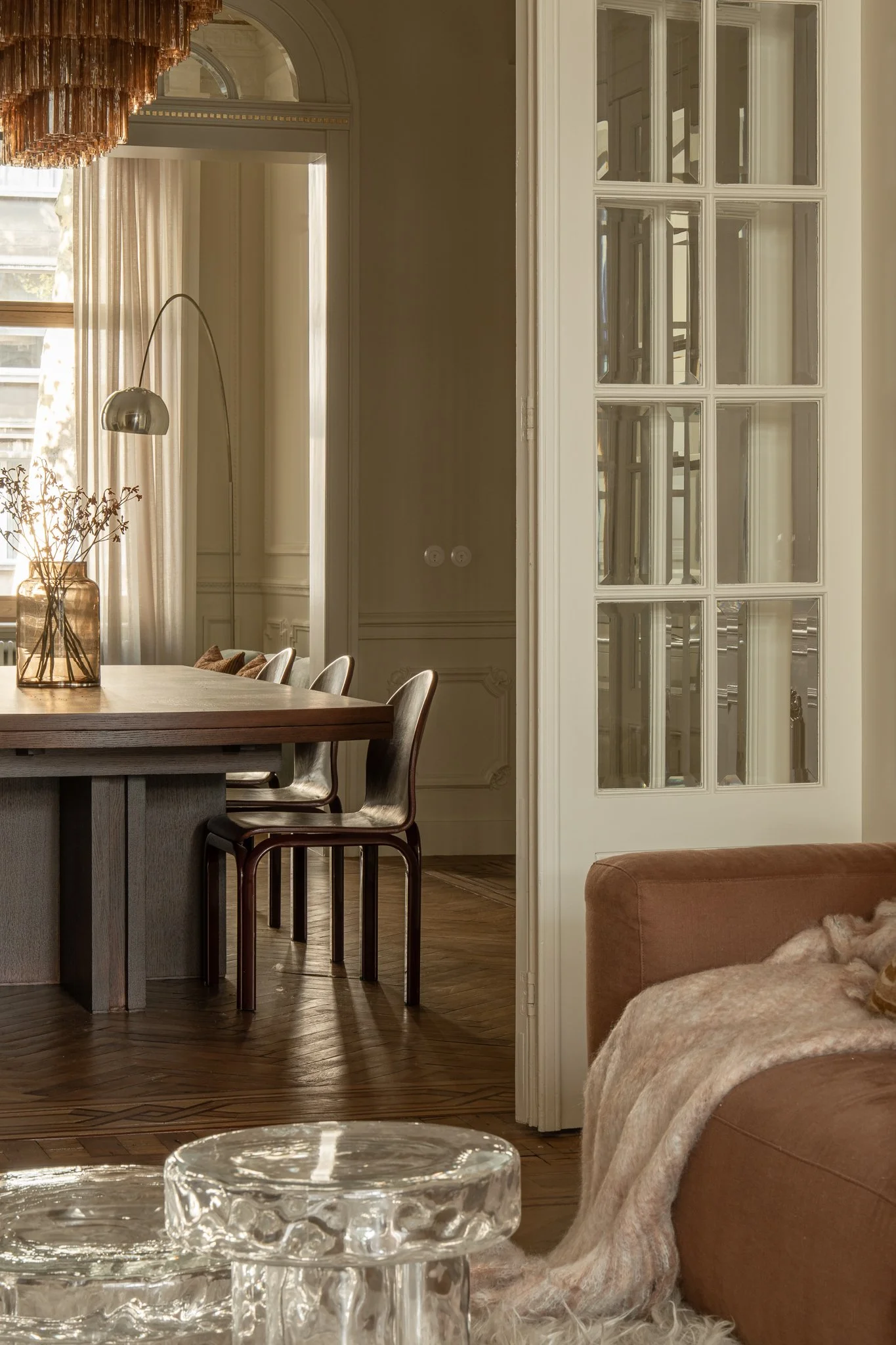A view of a dining room with a wooden table, chairs, a glass vase with dried flowers, and a window with curtains. Part of a sitting area with a tan sofa and a blanket is visible in the foreground.
