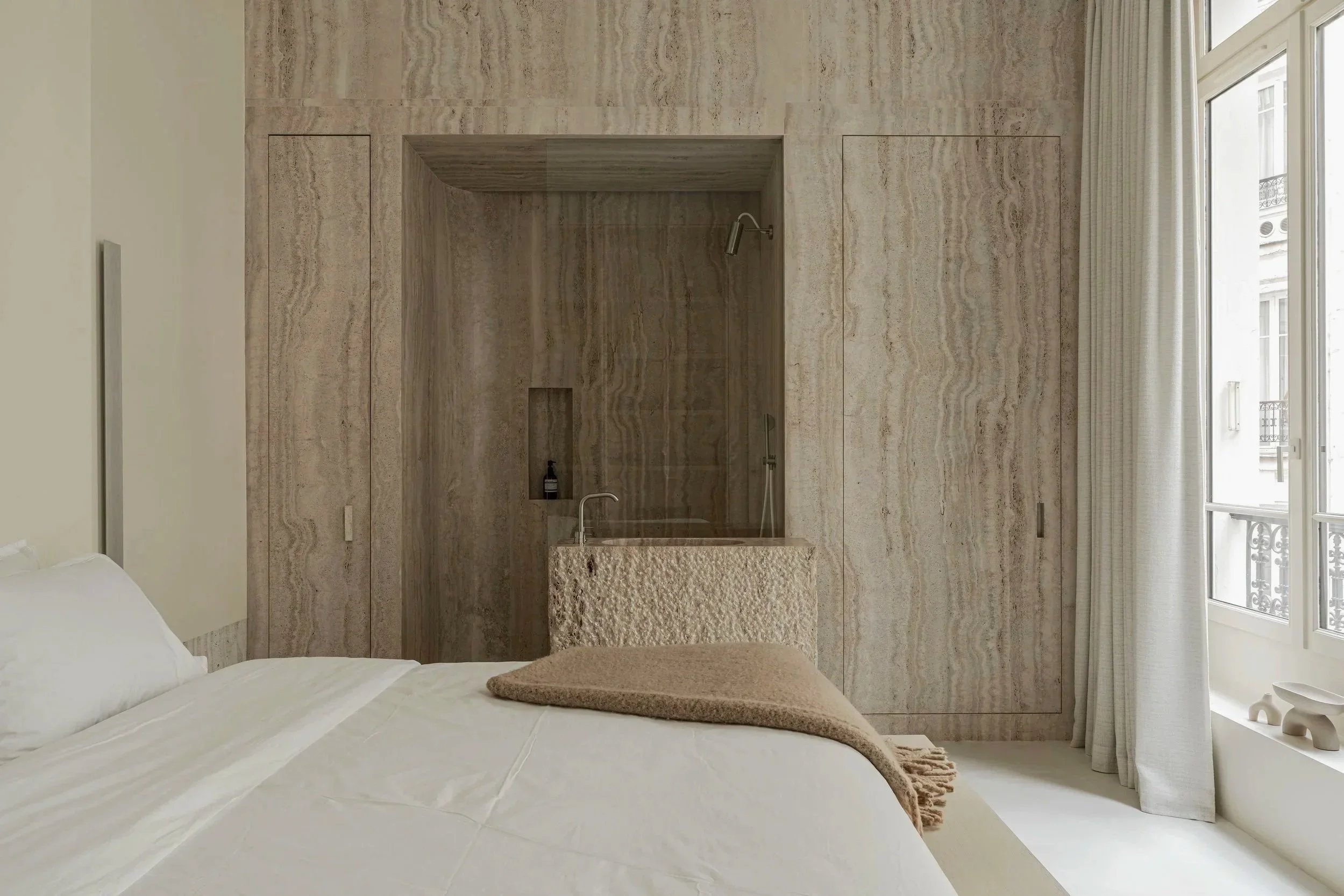 A bedroom with a bed, beige blanket, and a built-in shower area with a stone sink, marble walls, and large windows with white curtains.