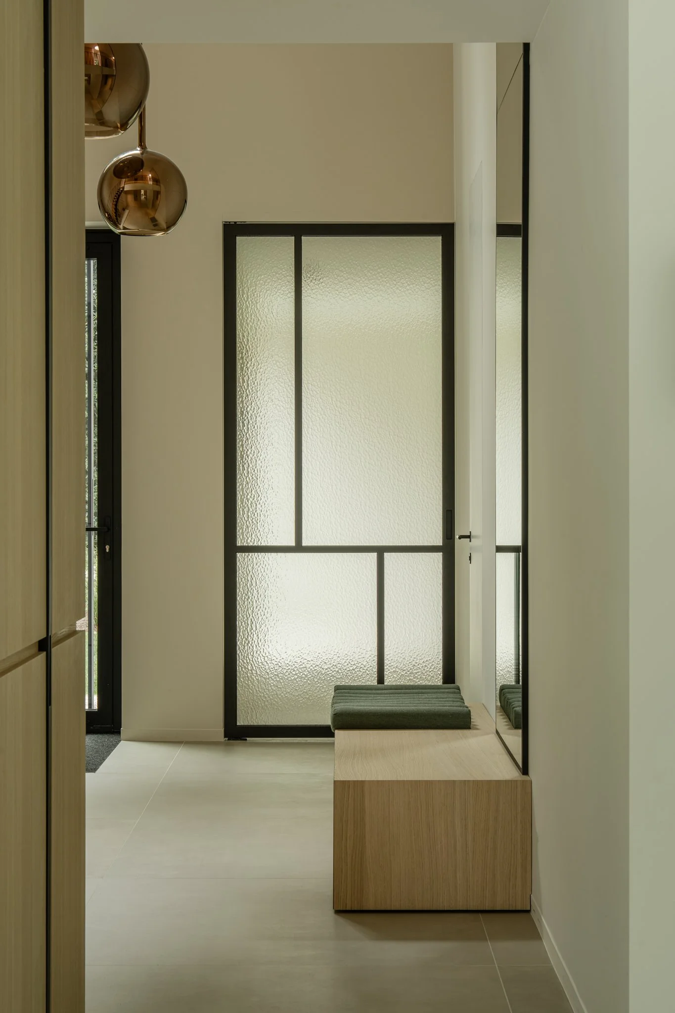 Interior view of a modern entryway or hallway with a frosted glass door, a wooden bench with green cushions, and a large mirror, with minimal decor.