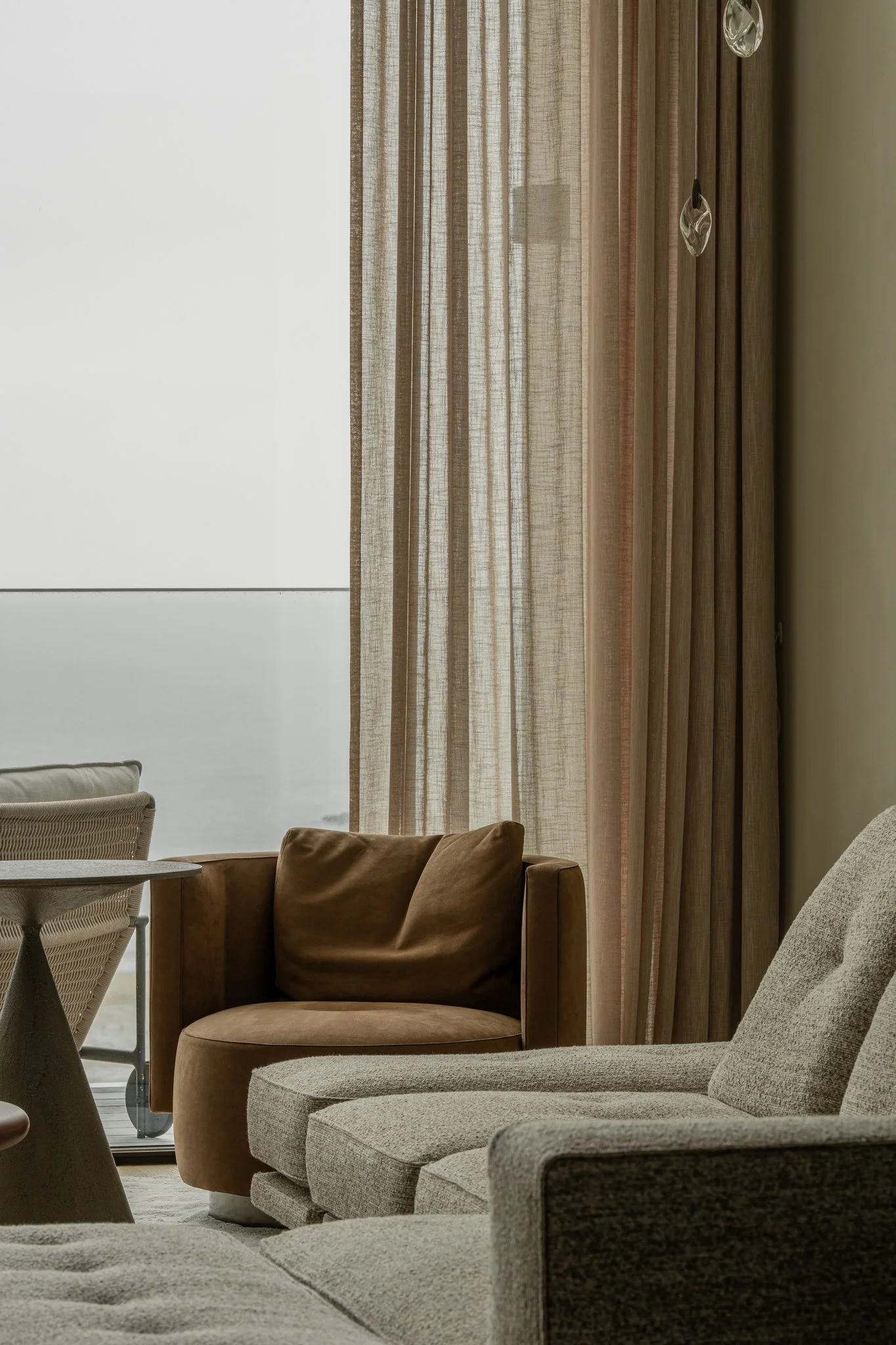 Living room with beige sofa, brown armchair, side table, and sheer curtains.