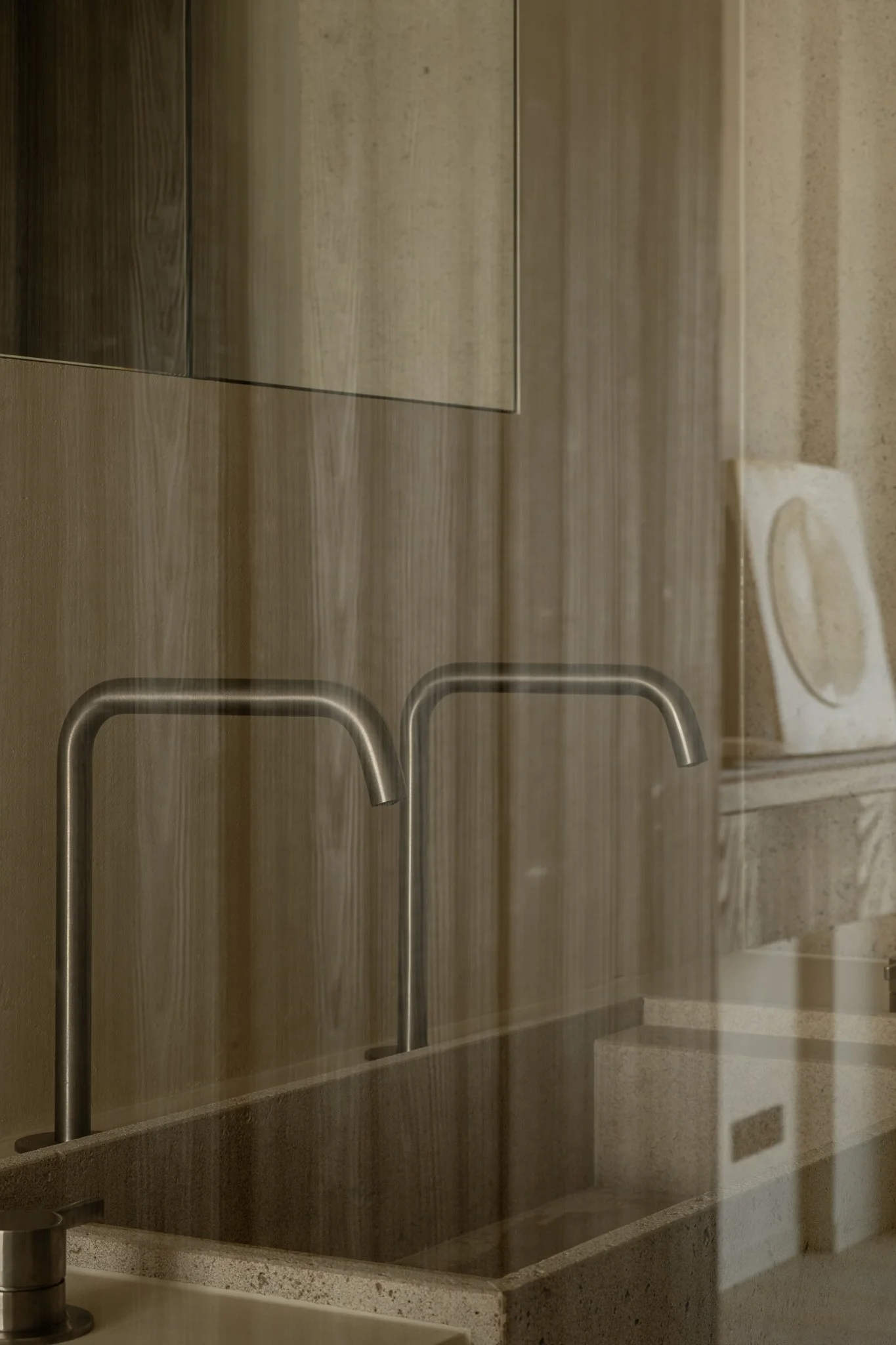 A double sink with chrome faucets in a modern bathroom with wood paneling and a mirror, reflected in a glass window or door.