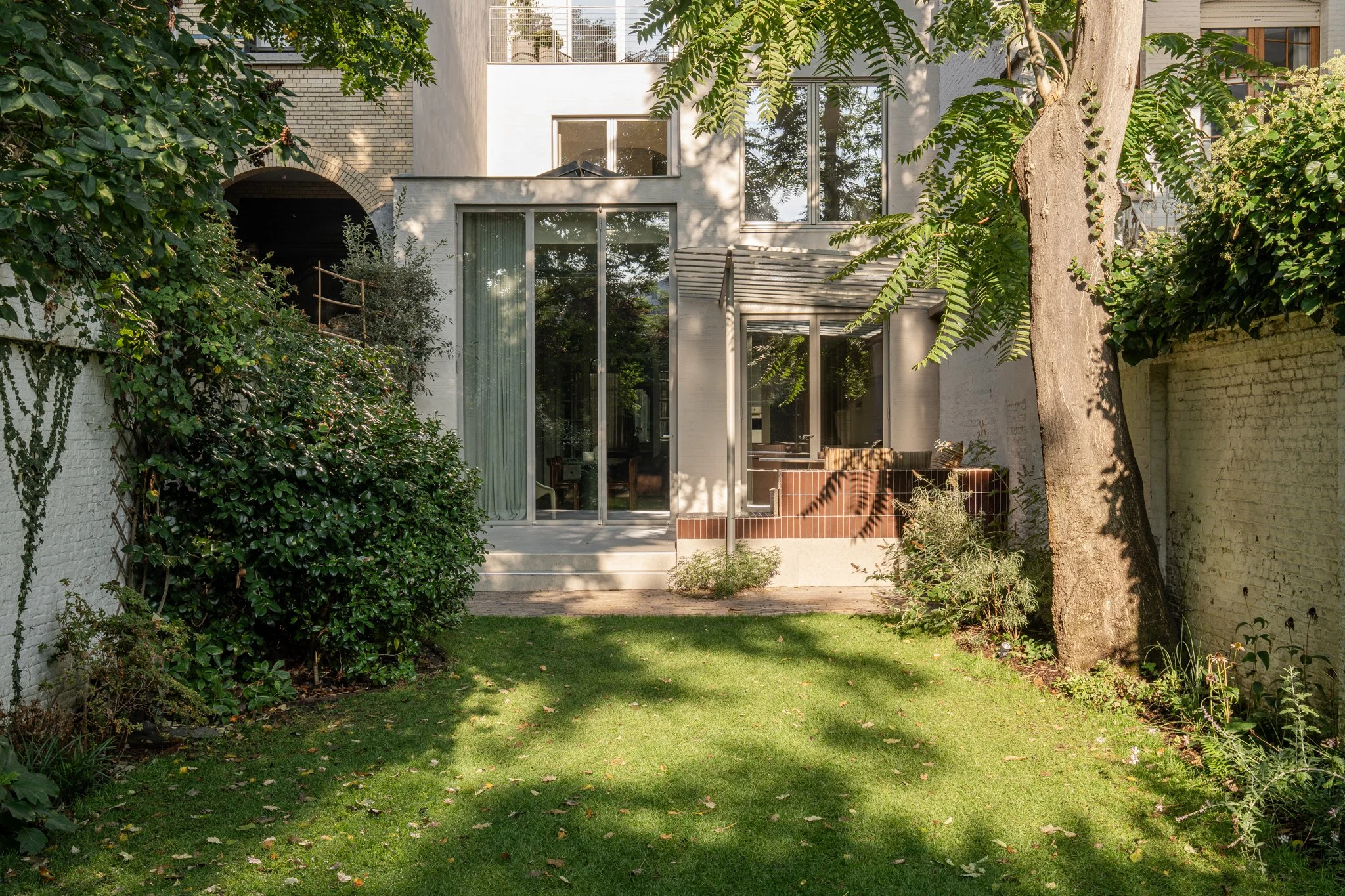 Backyard garden with green grass, bushes, trees, and a modern house with large glass windows and sliding doors.