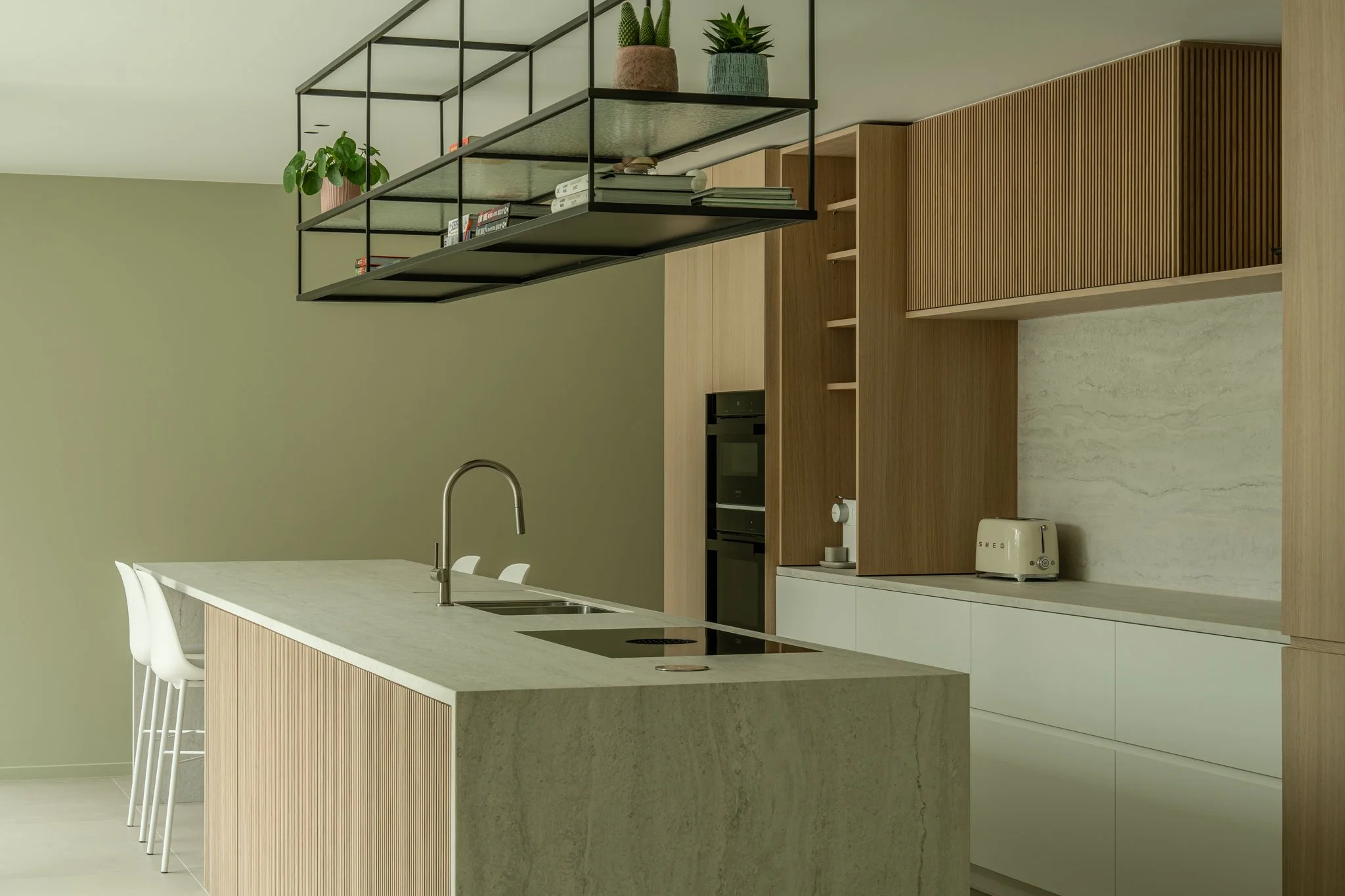Modern kitchen with a large island, white chairs, light wood cabinets, a built-in oven, a toaster, and a black metal hanging shelf with plants and books.