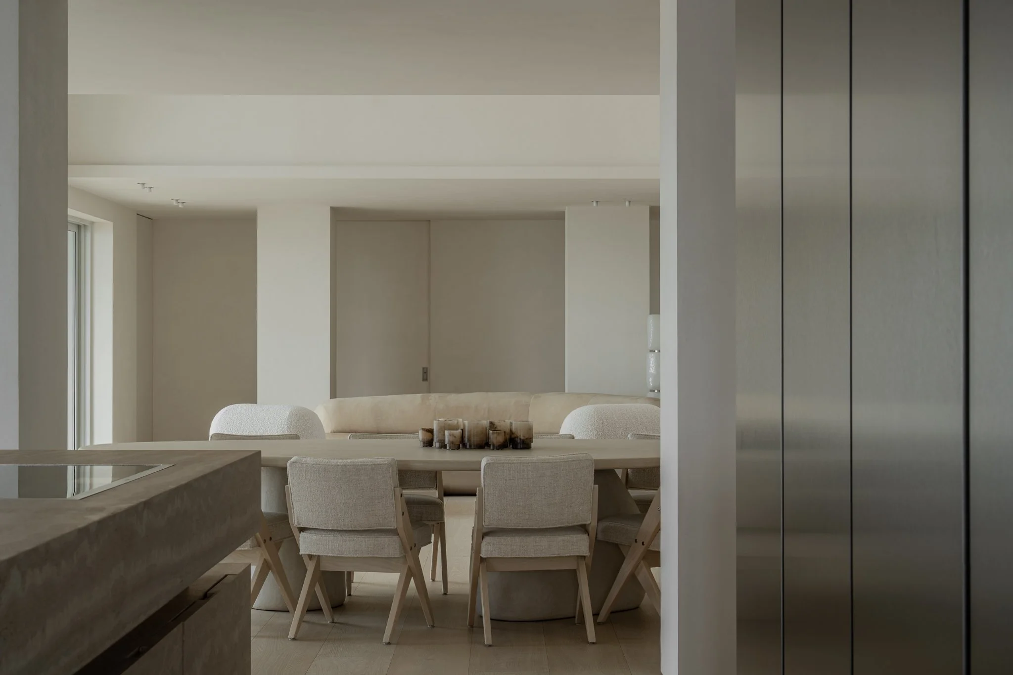 Minimalist, modern dining area with a round table surrounded by beige upholstered chairs in a neutral-toned room.