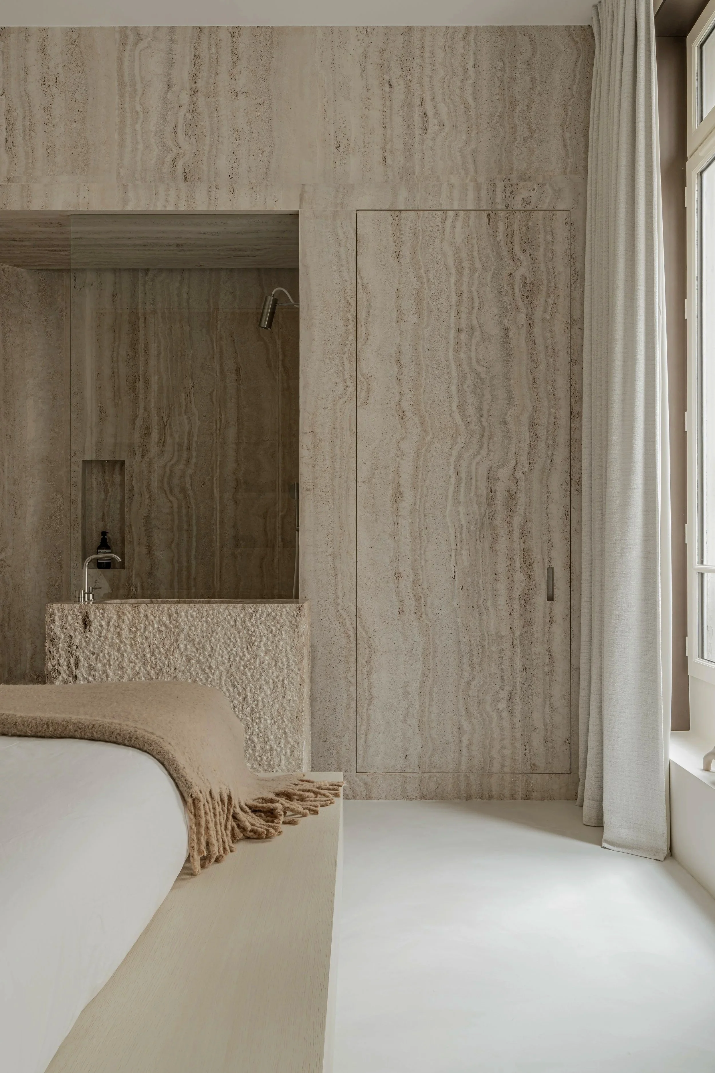 A minimalist bedroom with beige stone walls, a large window with white curtains, and a bed with a neutral-colored blanket partially visible in the corner.