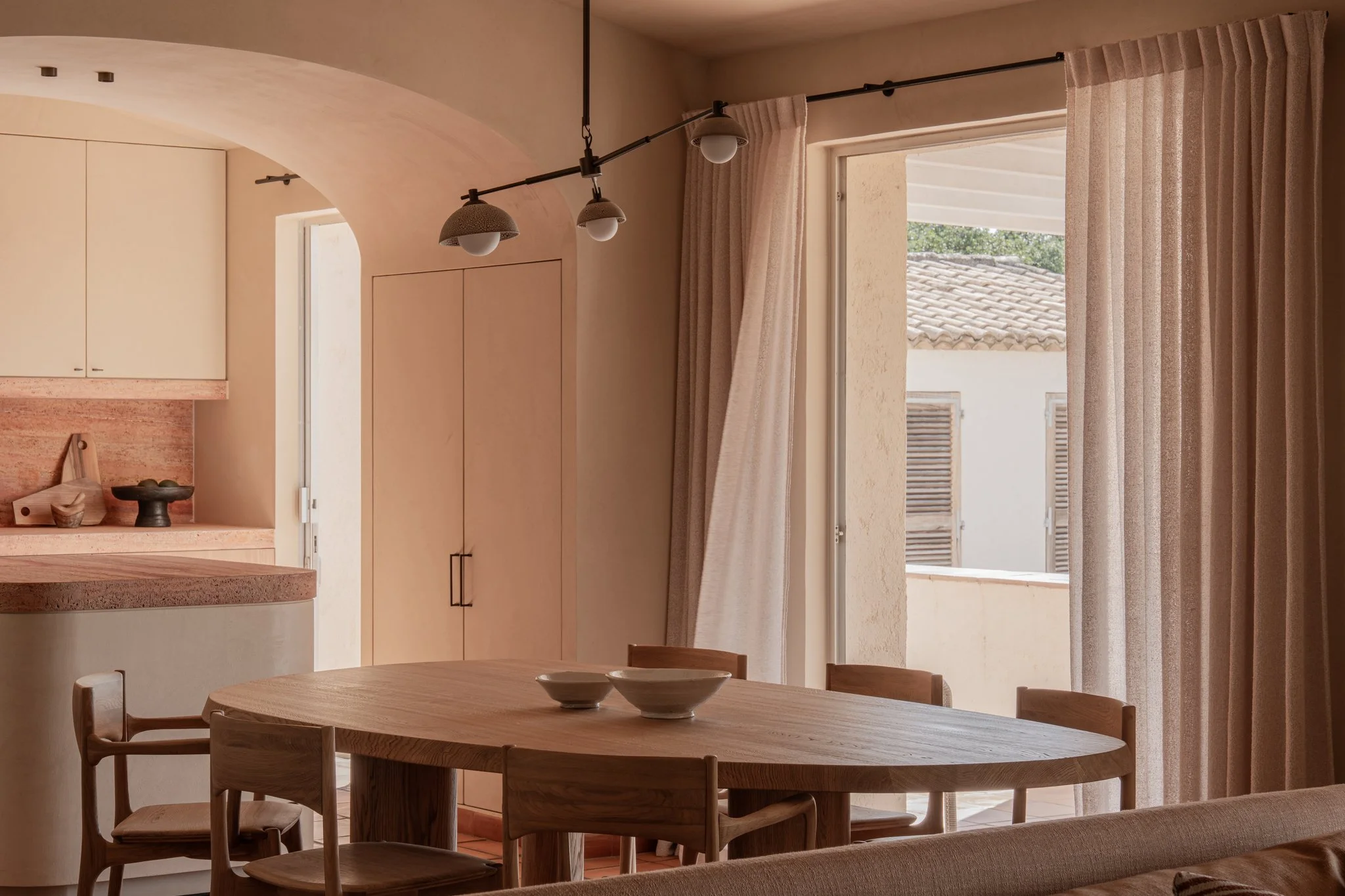 A cozy dining area with a wooden table and chairs next to large windows with beige curtains, overlooking neighboring houses with tiled roofs.