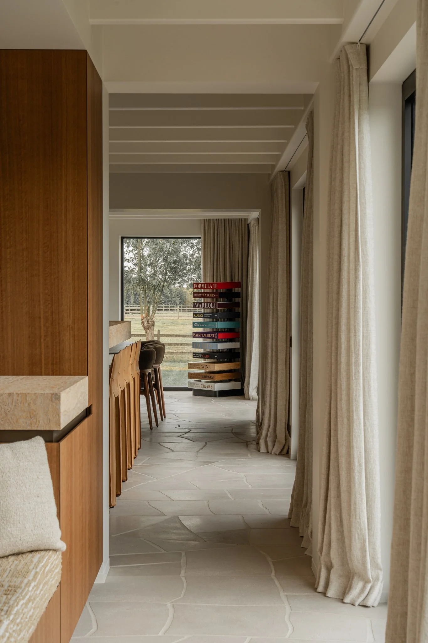 Interior view of a modern room with beige curtains, a wooden kitchen counter, and high chairs, leading to a window and a stack of books or magazines on a black spiral stand by the glass door.