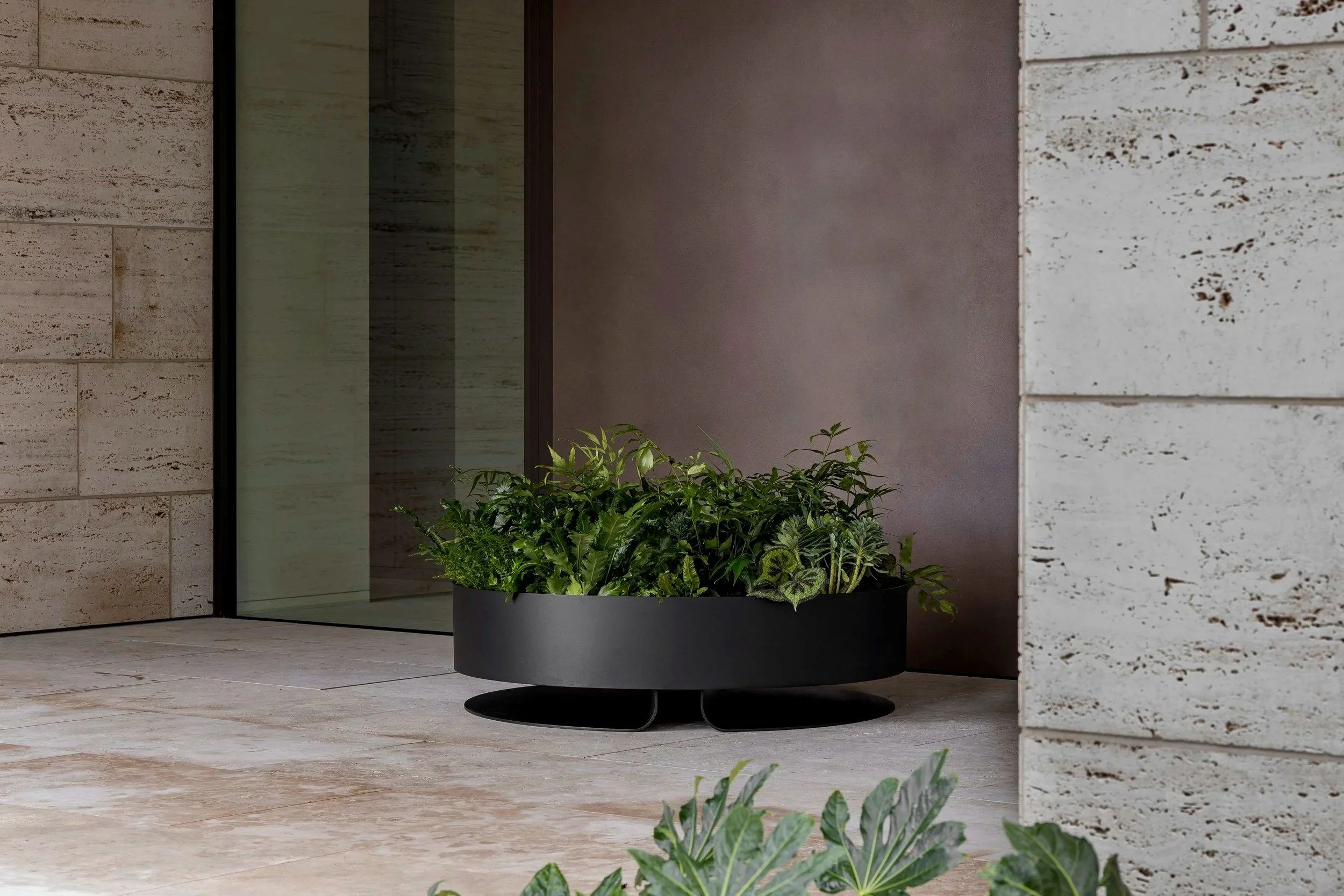 A modern outdoor space with a large black planter filled with various green plants, positioned next to a glass door and surrounded by beige and white stone walls.