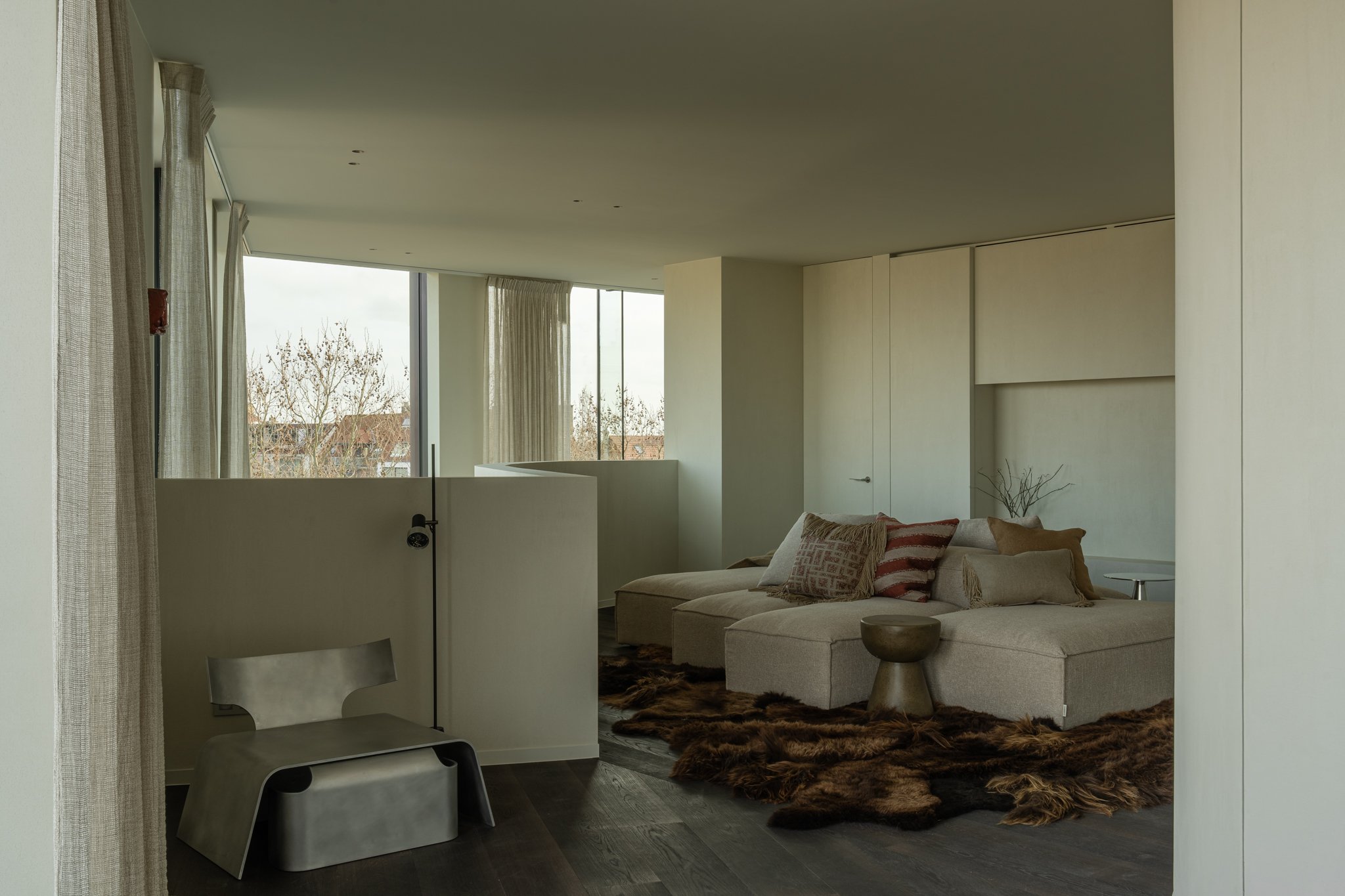 Modern living room with beige sectional sofa, decorative pillows, a small side table, and a fur rug. Large windows with cream curtains allowing natural light to fill the space.