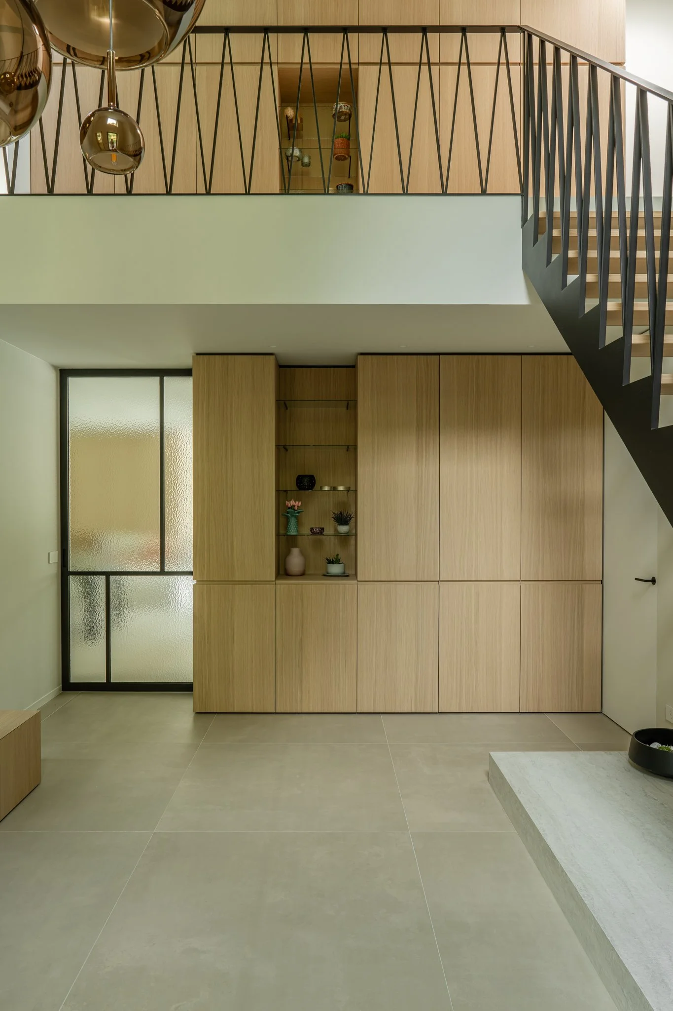 Interior of a modern home with wooden cabinets, frosted glass sliding door, staircase with black railing, and beige tile flooring.