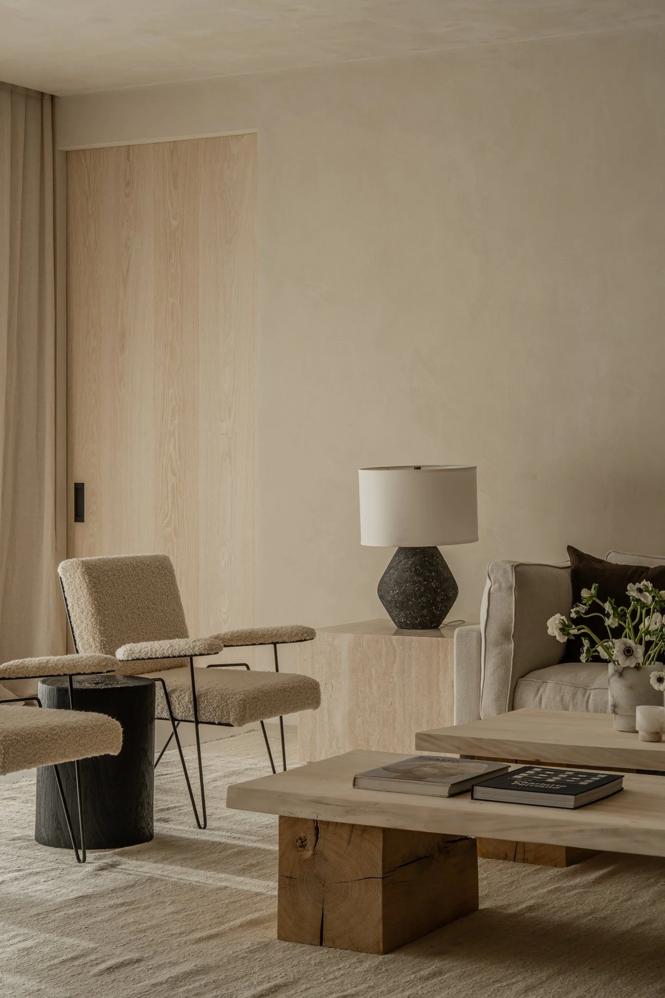 Living room with beige walls, a wooden door, a beige sofa with pillow, a black and white table lamp, a coffee table with books, and a sitting chair with a side table.