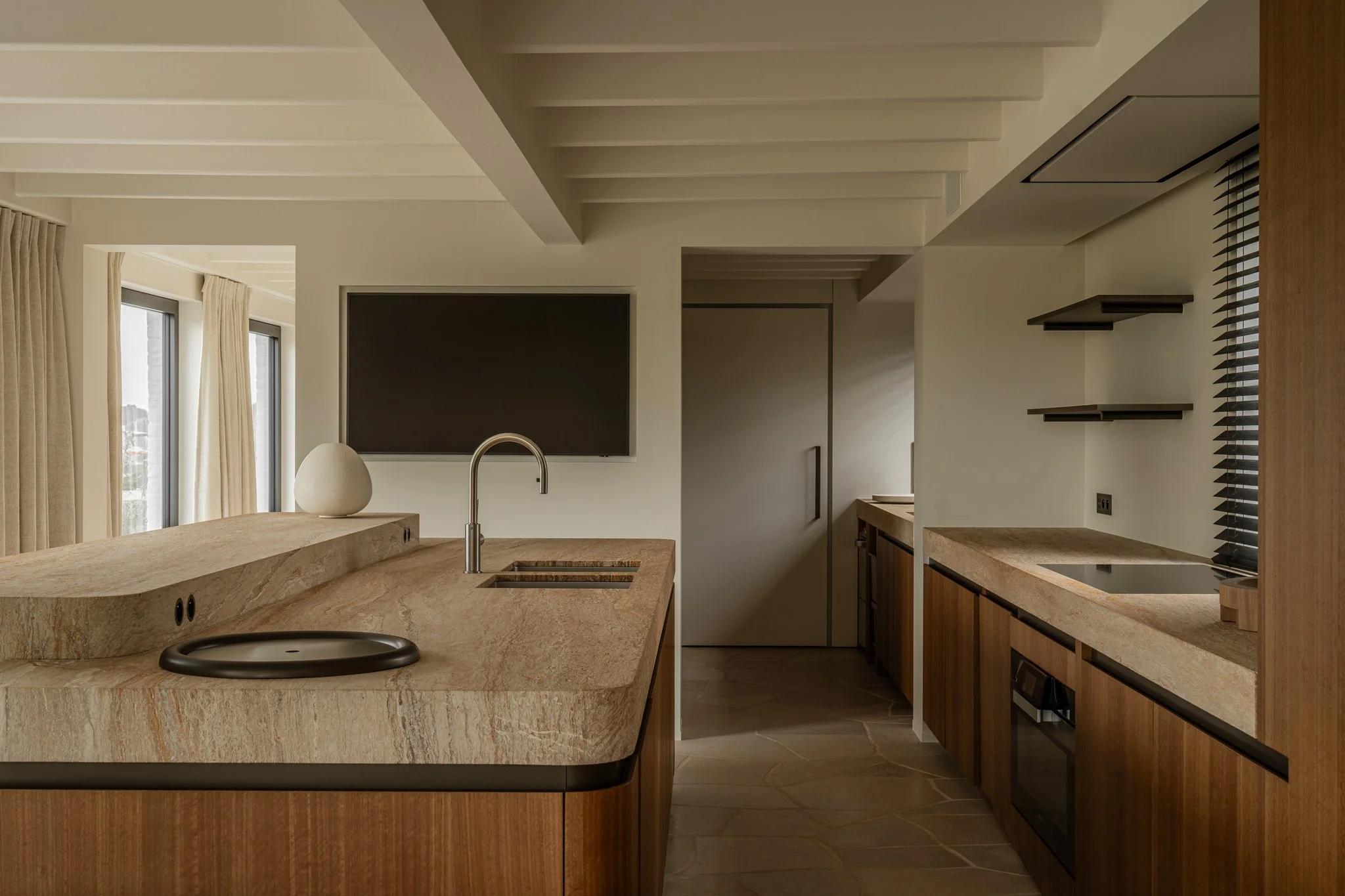 Modern kitchen with beige stone countertops, wooden cabinets, black stovetop, and a wall-mounted TV, with large windows and curtains in the background.