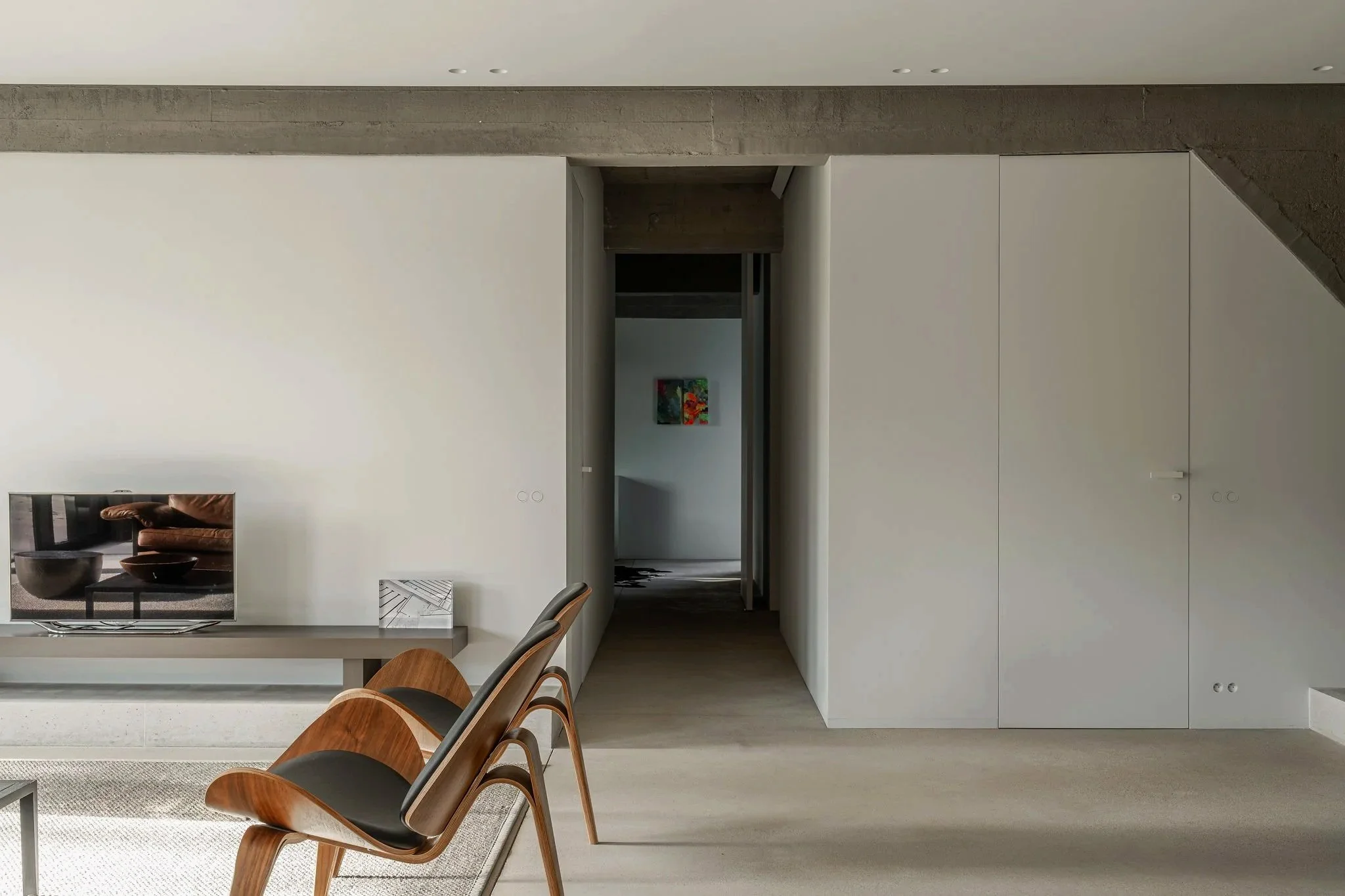 Interior of a modern minimalist living space with white walls, a flat-screen TV on a gray console, wooden chairs with black seats, and an open doorway leading to another room with artwork on the wall.