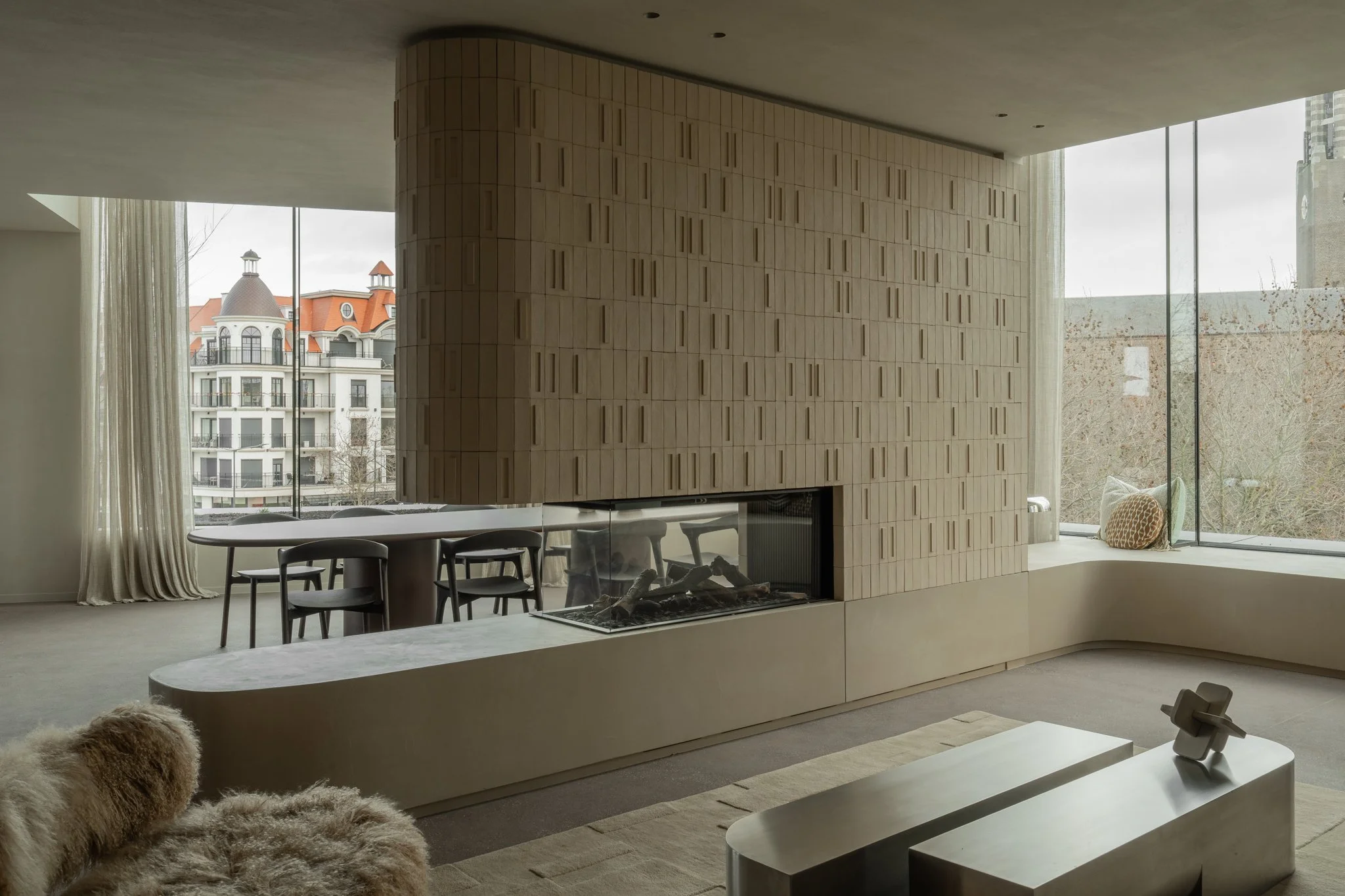 Modern living room with large windows, a built-in fireplace, a white coffee table, and a beige sofa with furry cushions, overlooking buildings with red roofs.