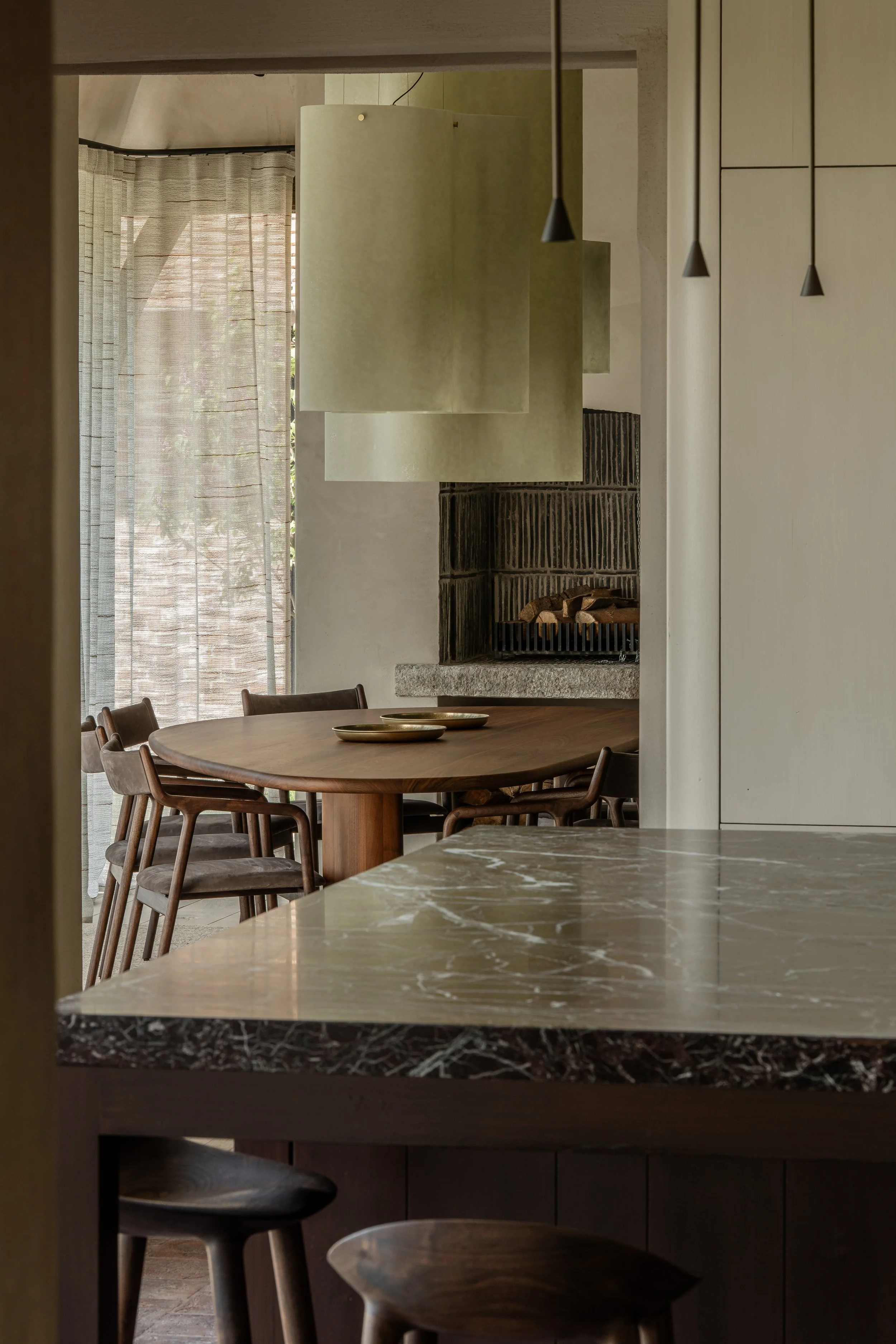 A dining area with a round wooden table, chairs, a brick fireplace, and light curtains.