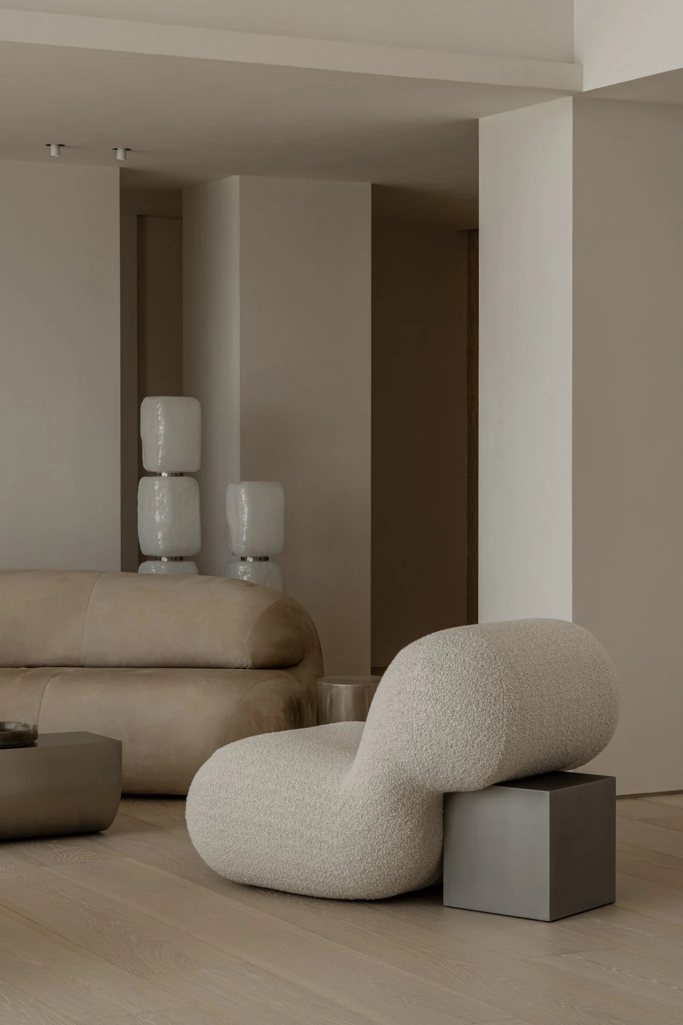 Modern minimalist living room with beige and cream furniture and decor, including a plush curved armchair, a beige sofa, and abstract vases.