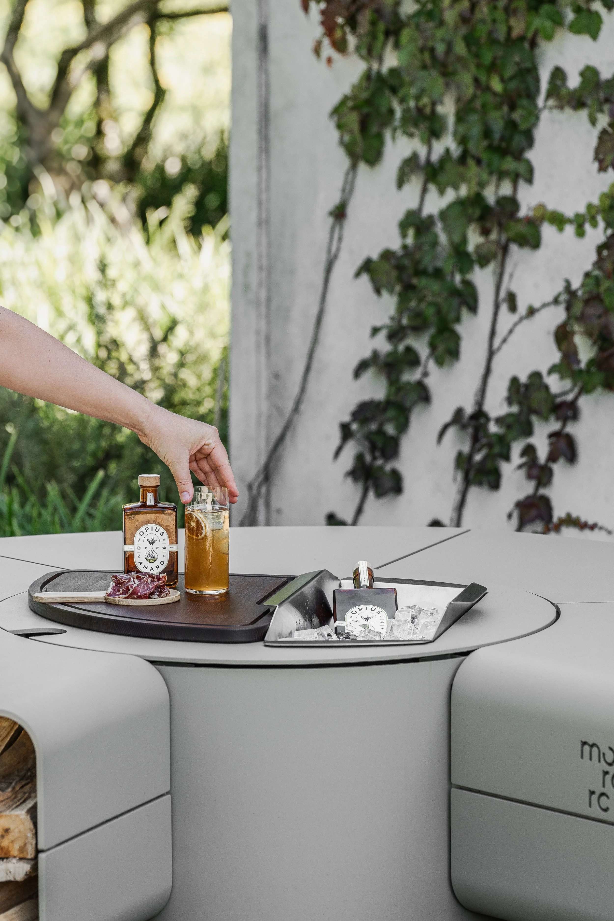 Person's hand reaching for a glass of iced tea with lemon and a bottle of Opus Amaro liquor on a black tray, alongside a piece of raw meat, on a white outdoor table with greenery in the background.