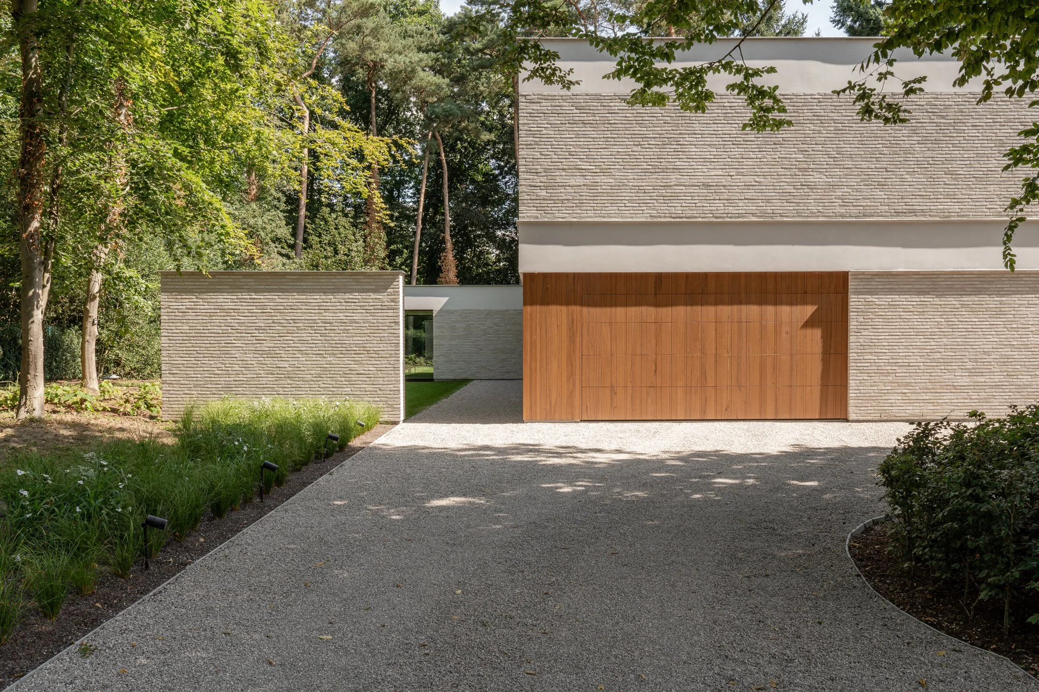 Modern house with light brick and wooden garage door, surrounded by trees and landscaped garden with gravel driveway.