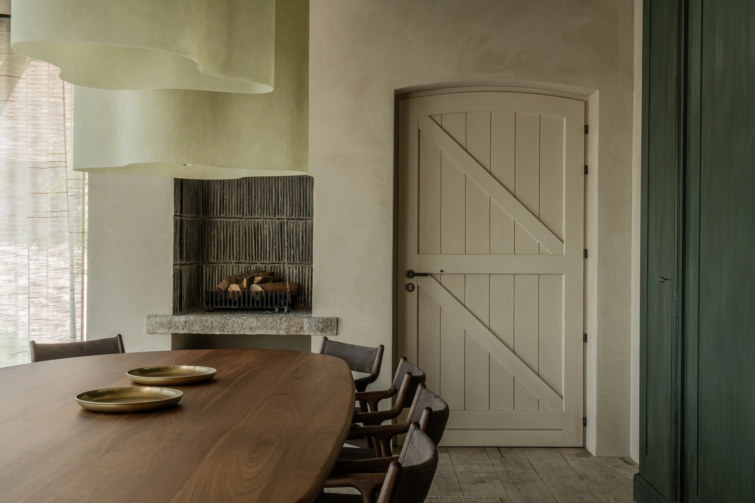 Interior of a dining room with a wooden table, four chairs, a stone fireplace, a closed wooden door, green cabinets, and hanging green lamp shades.
