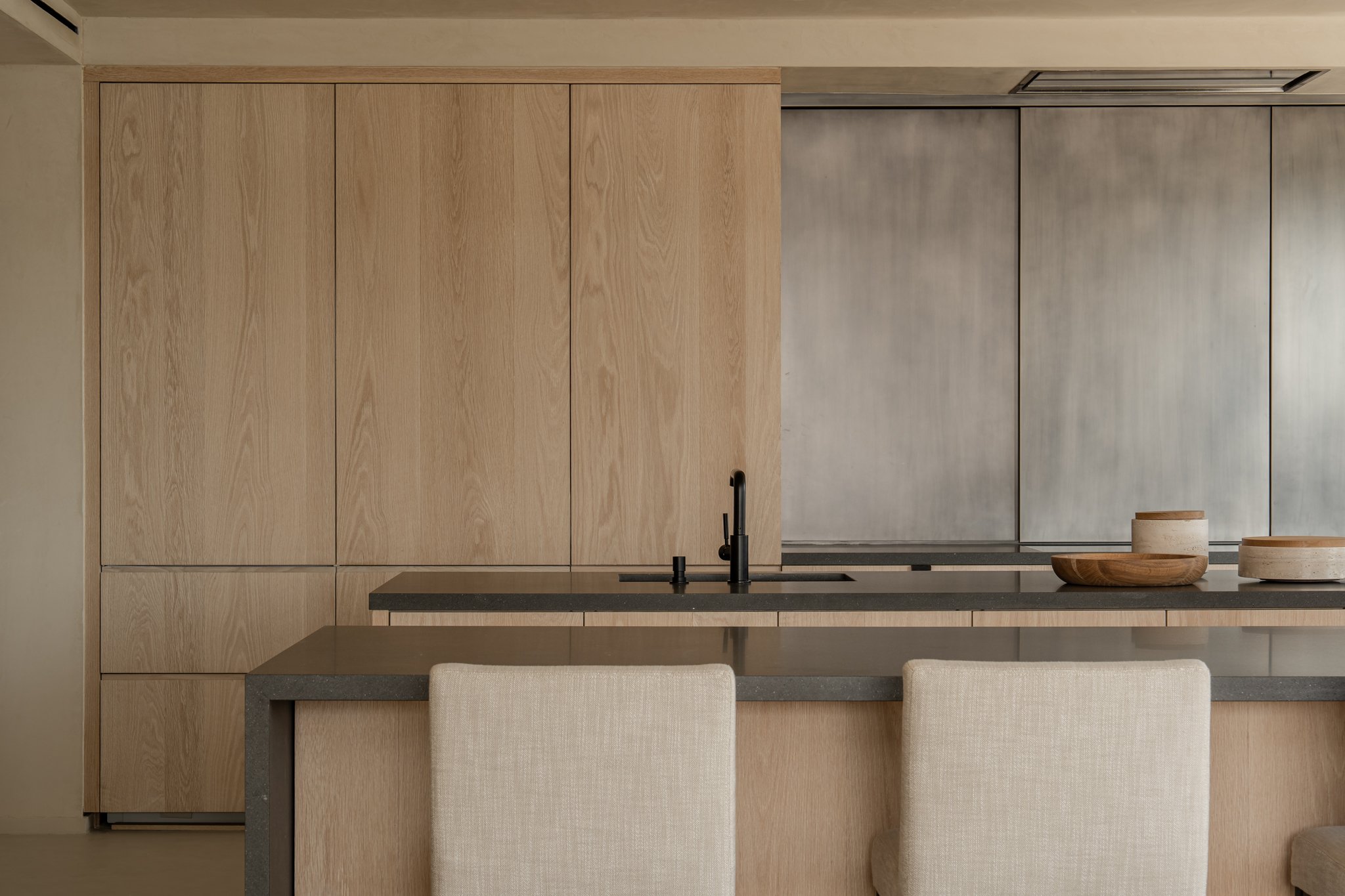 Modern kitchen with light wood cabinets, a black faucet, and a dark gray countertop. Two wooden bowls and a ceramic container are on the counter. Beige upholstered chairs are in front of the island.