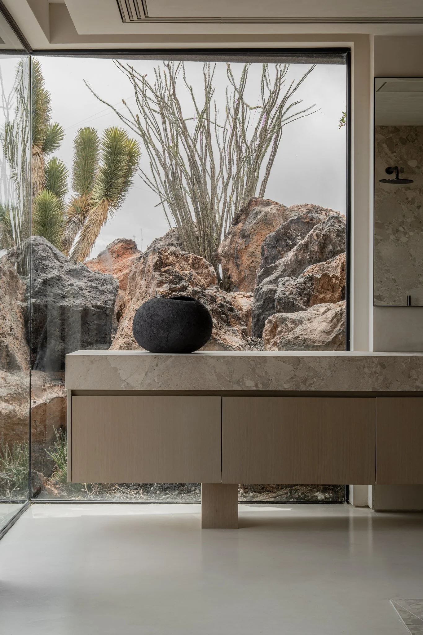 Modern bathroom with a large window showcasing desert rocks and plants outside. Contains a light-colored wooden vanity with a marble top and a round black stone object.