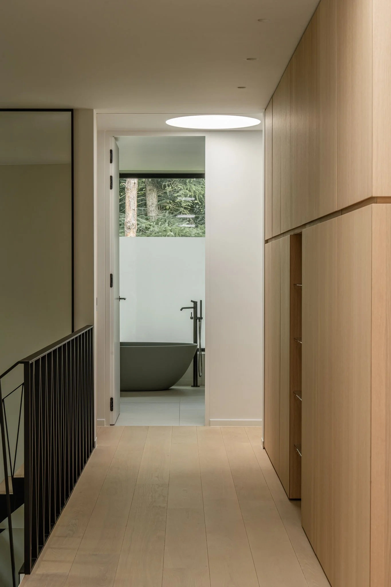 An interior hallway leading to a bathroom with a freestanding bathtub, seen through a door. The hallway has light wood flooring and built-in wooden cabinets along the right wall.