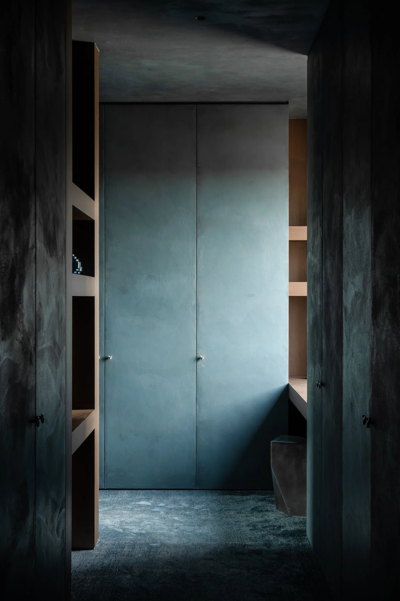 A narrow walk-in closet with tall dark shelves on each side, and a blue wardrobe with two knobs in the center background. The space has a textured, dark carpet and minimal lighting.