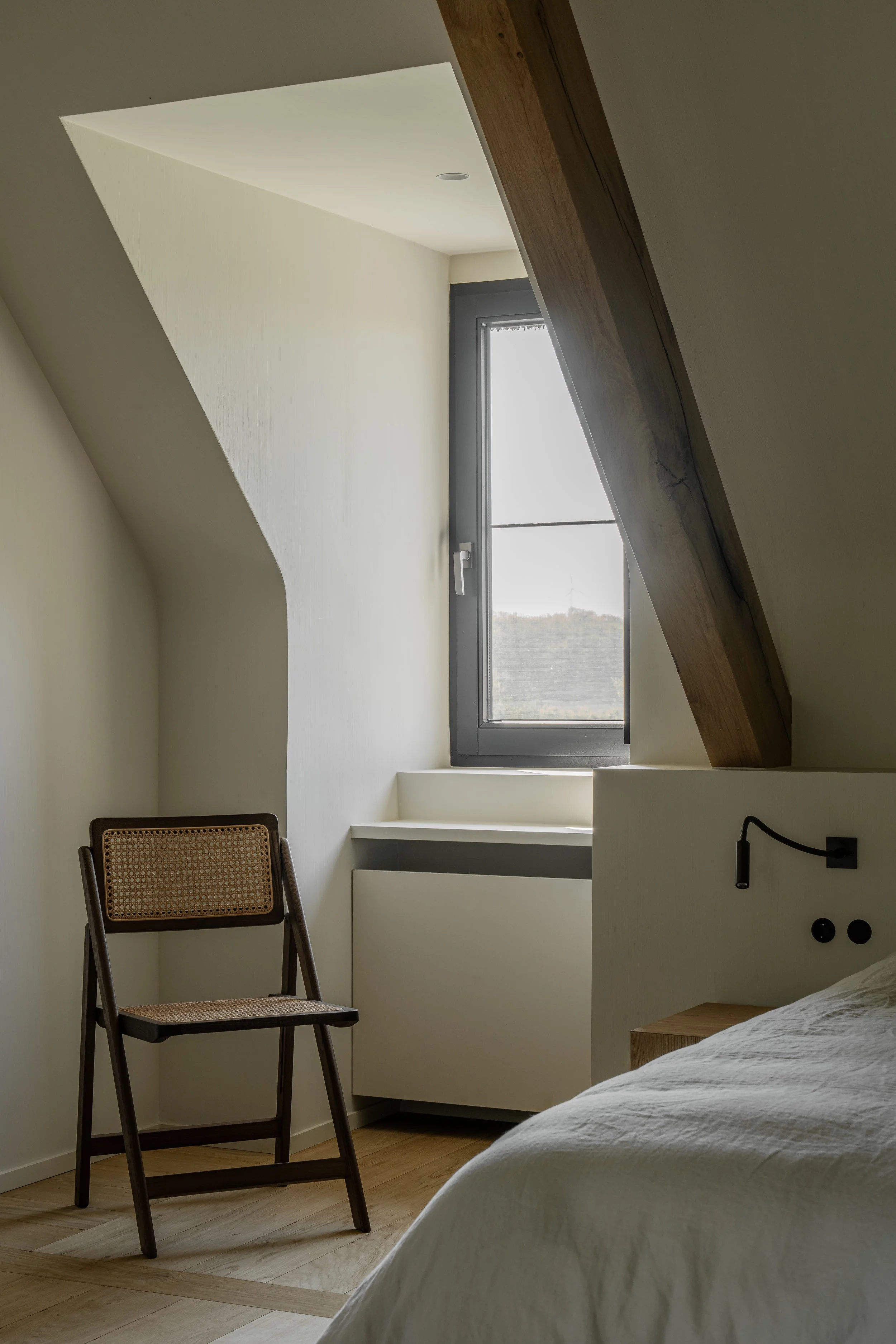 Interior view of a minimalist bedroom with a bed, a wooden chair, a window, and unique architectural ceiling details.