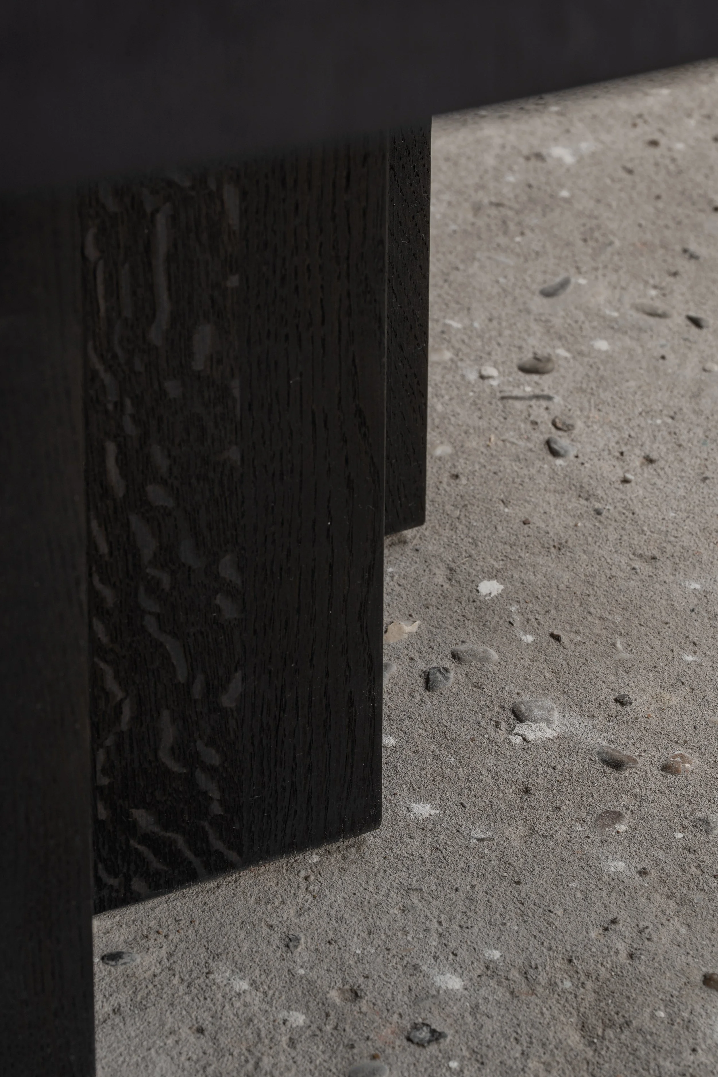Close-up of a black wooden table leg on a sandy surface with small pebbles.