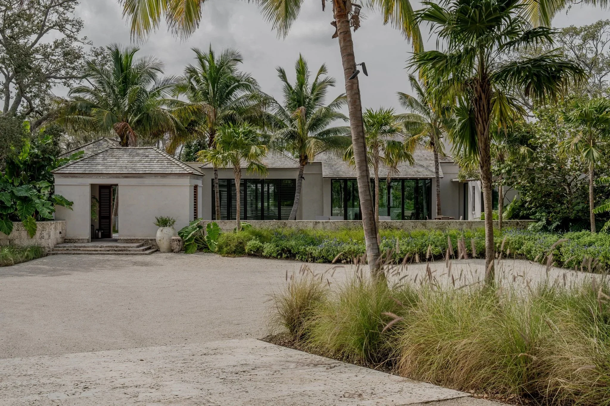 A modern house surrounded by palm trees and sandy landscaping, with a gravel driveway and lush greenery.