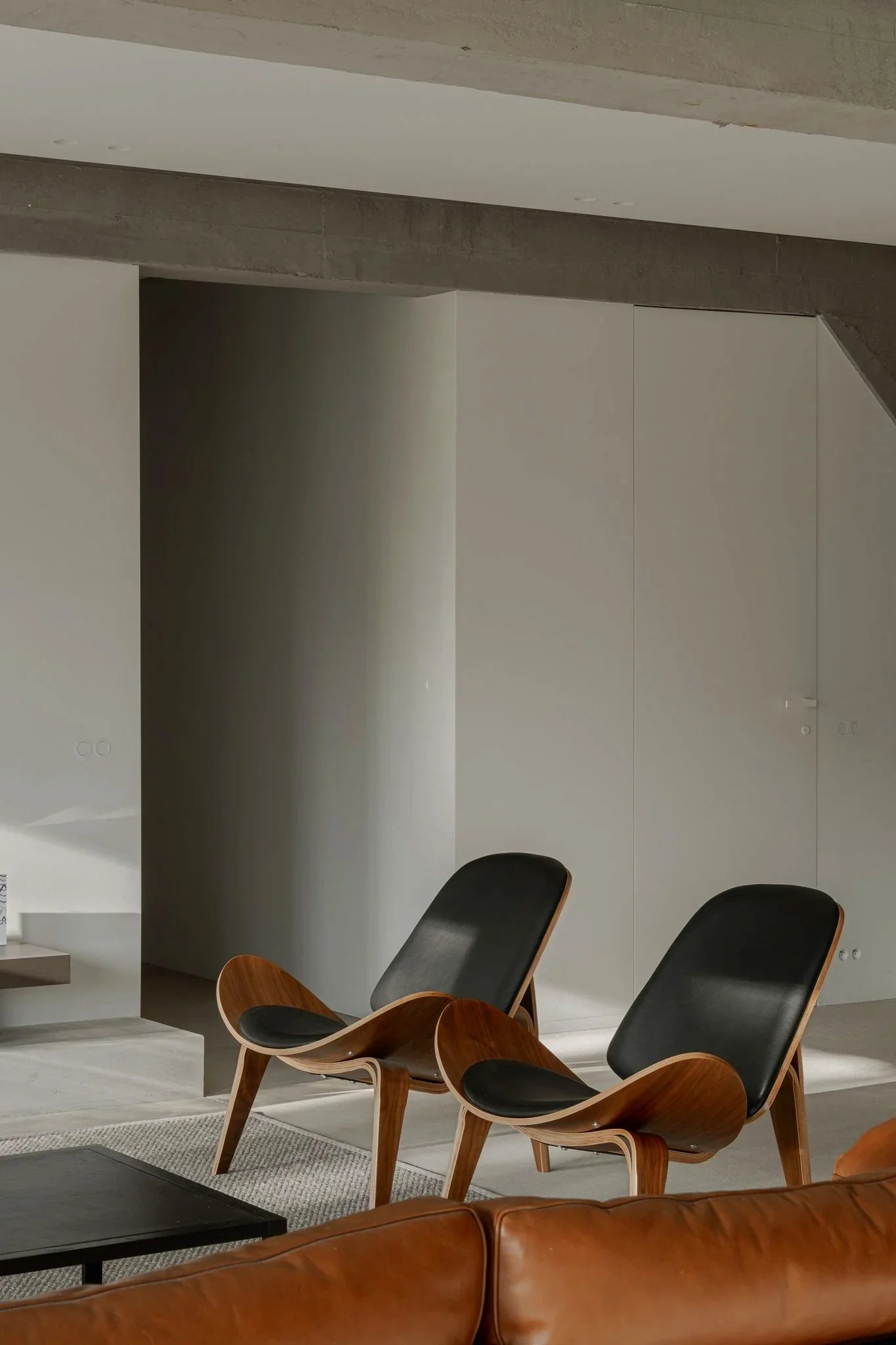 Two modern lounge chairs with black leather seats and wooden frames are positioned in a minimalist interior space with light gray walls and a concrete ceiling. A brown leather couch is partially visible in the foreground.