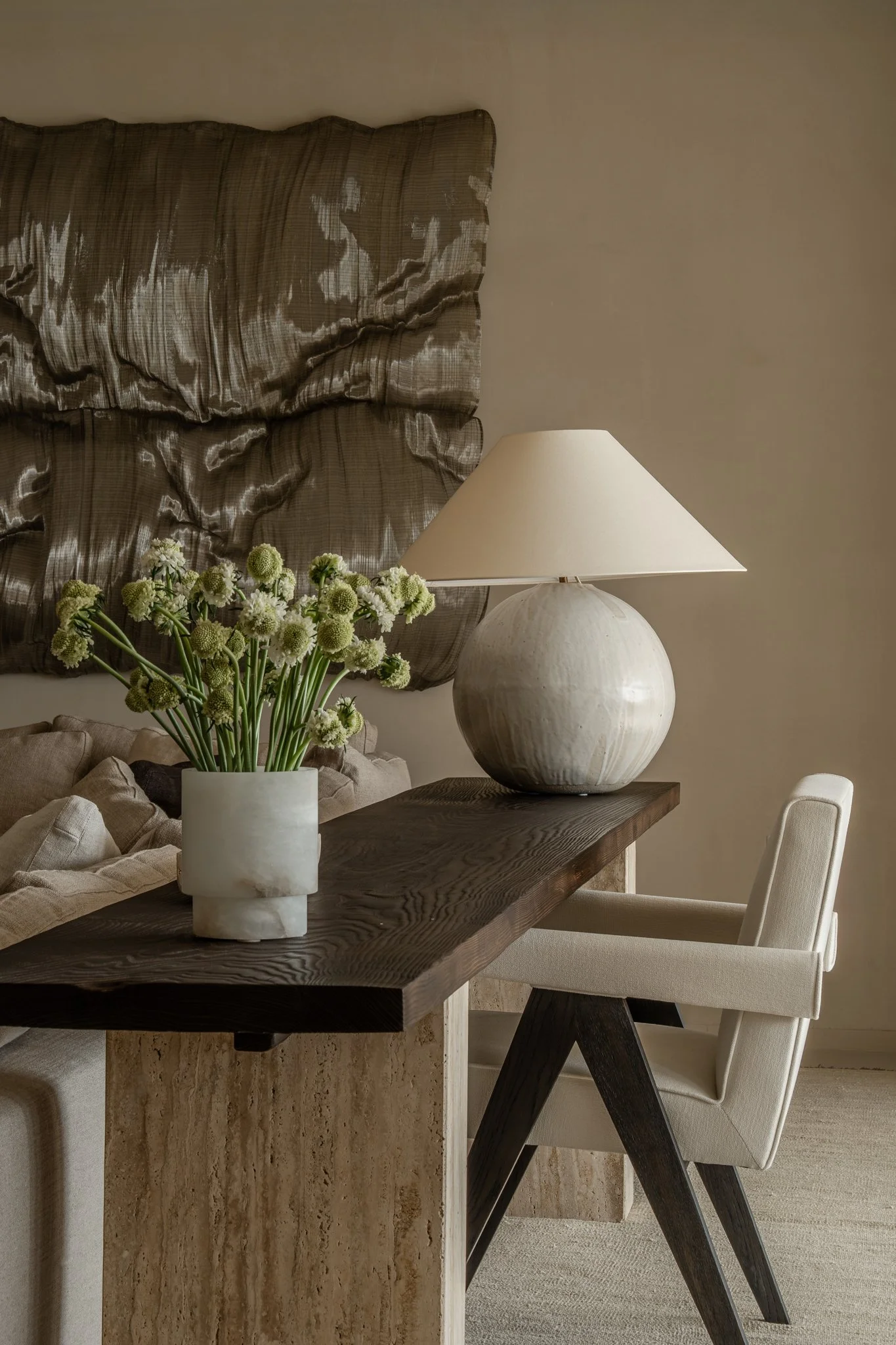 A living room corner featuring a wooden table with a white vase of green and white flowers, a large ceramic table lamp, a beige armchair, a sofa with cushions, and a textured fabric wall hanging.