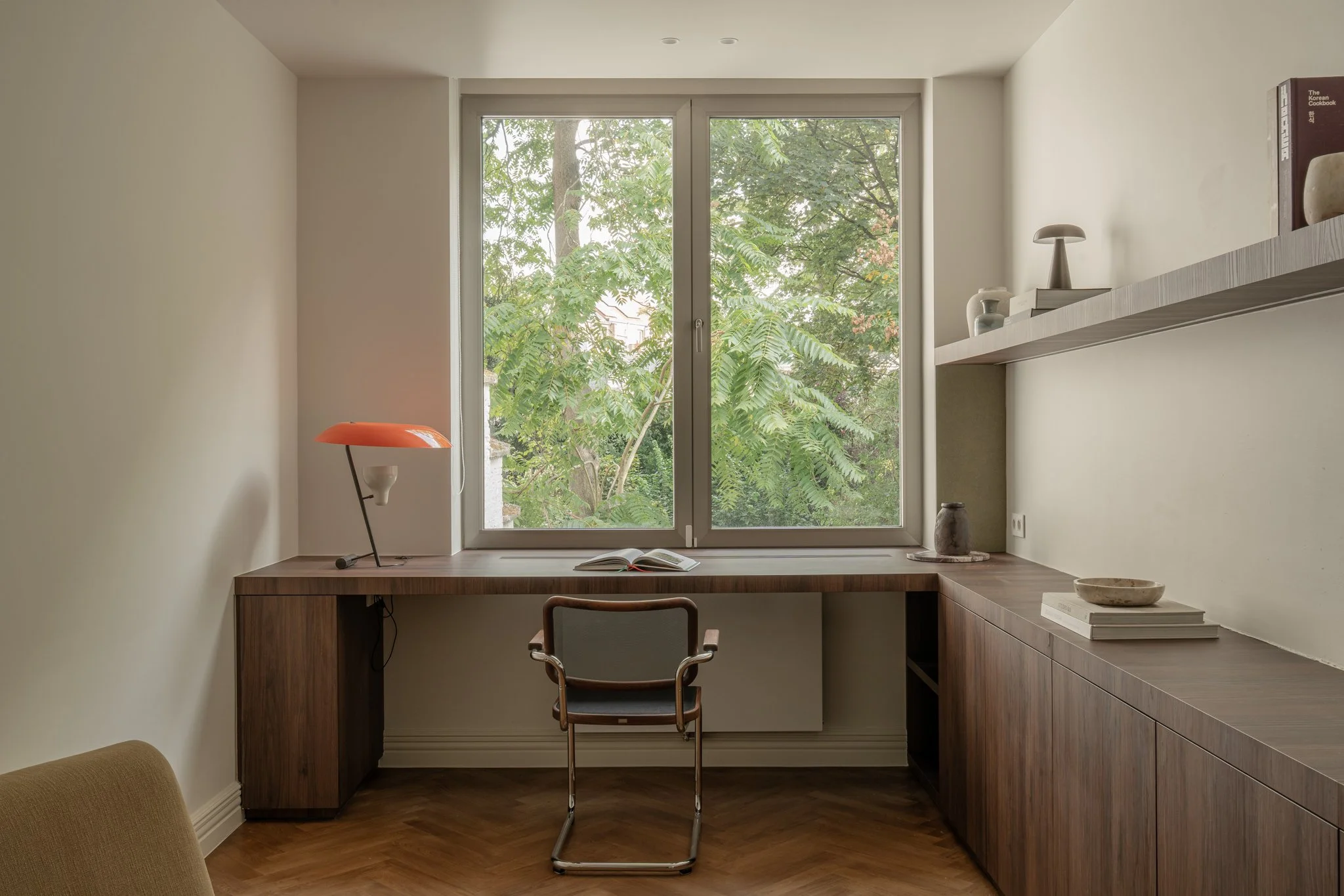 Home office with wooden desk, window overlooking green trees, modern chair, orange desk lamp, open book, decorative items on wall shelf, and neutral wall colors.