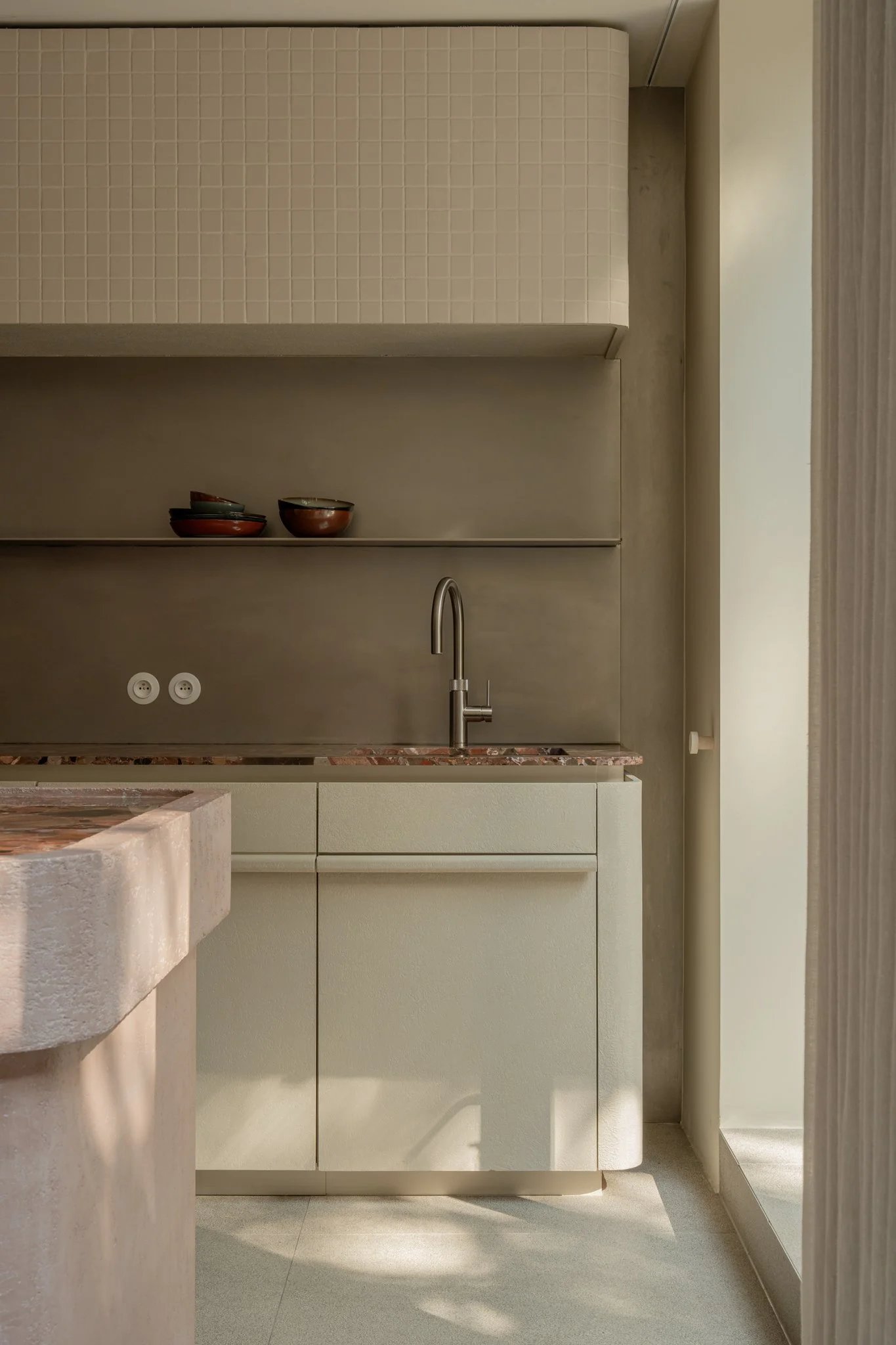 Minimalist kitchen with beige cabinets, a marble countertop, and a stainless steel faucet above the sink. Open shelves with bowls and a textured wall panel.
