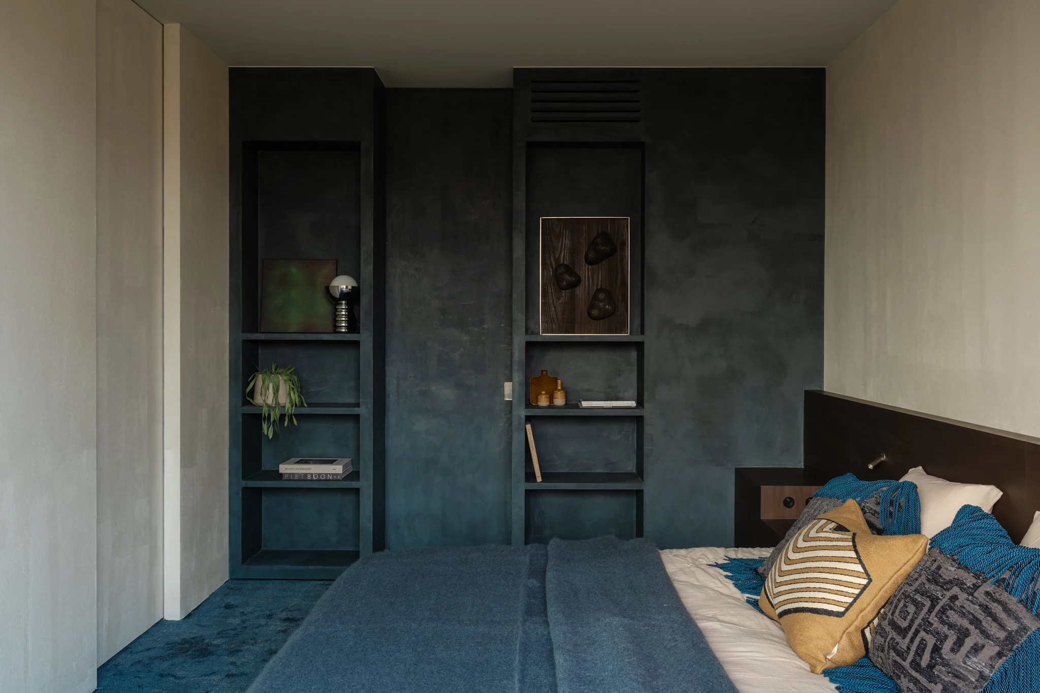 A bedroom with a dark accent wall, built-in shelves, a bed with gray, beige, blue, and patterned pillows, and a dark headboard.