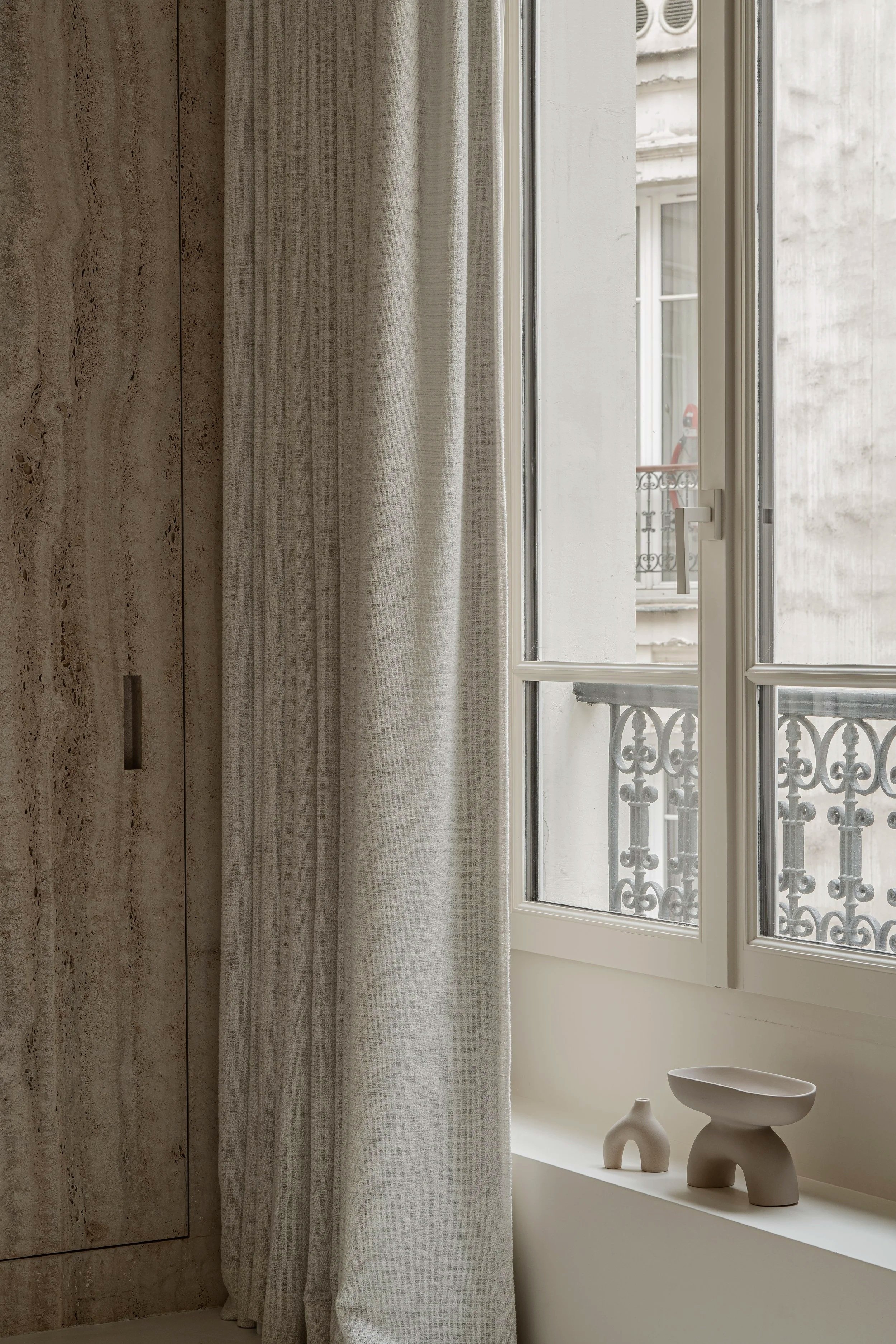 A window with a beige curtain, decorative ceramic objects on the windowsill, and a view of a neighboring building with wrought iron balcony railings.