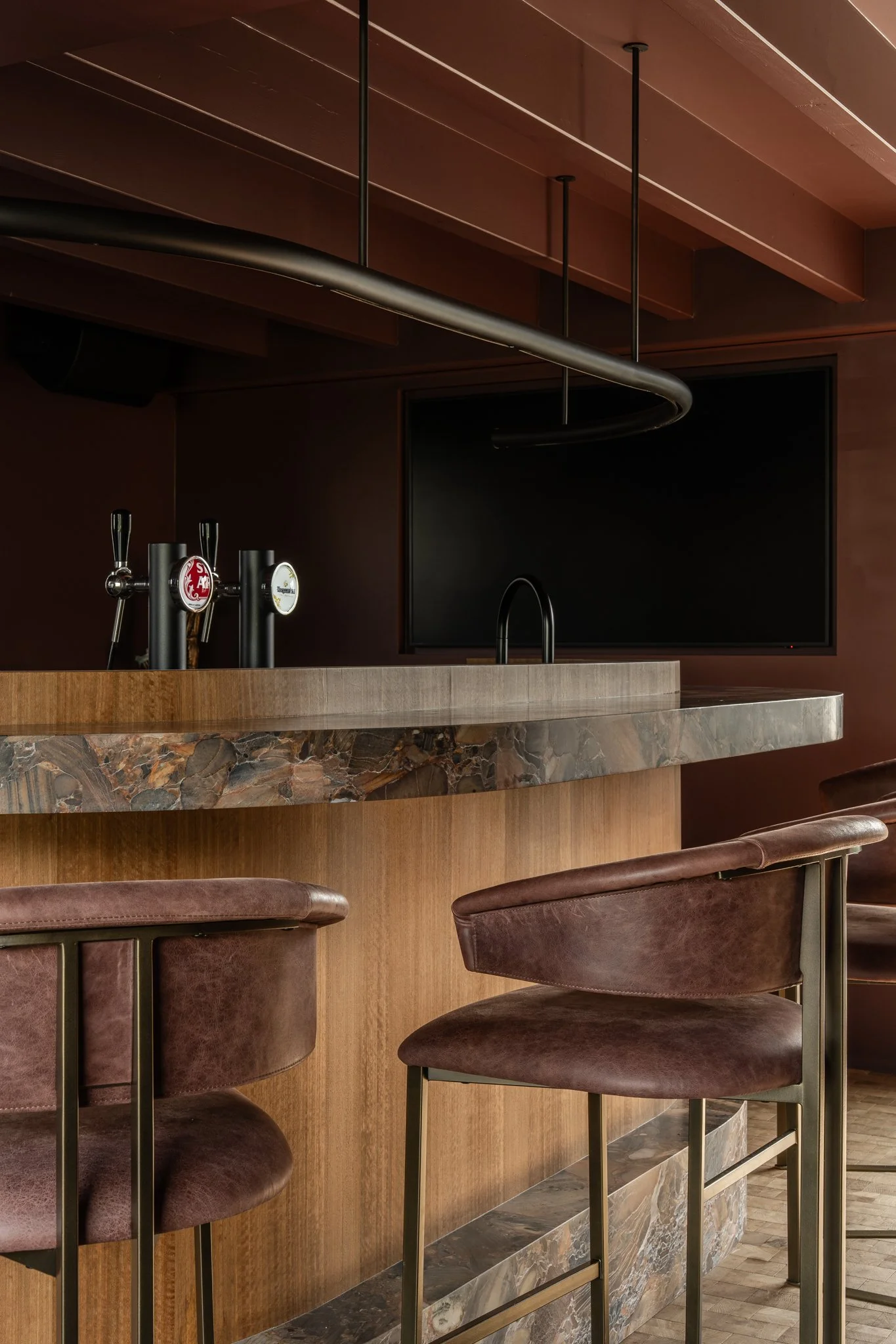 A modern bar with a marble countertop, leather bar stools, a large black television, and beer taps, set against dark wood walls.