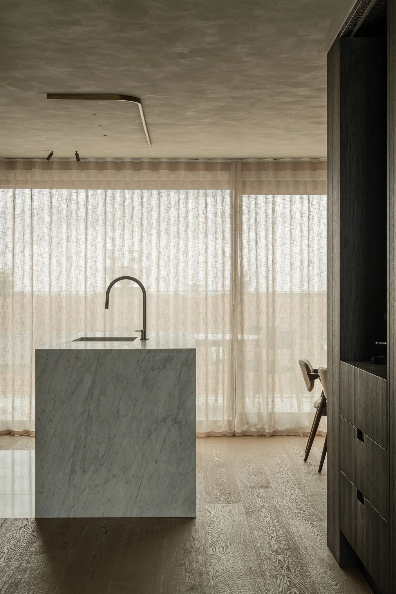 Minimalist kitchen with a marble island, a modern faucet, light wooden floor, and sheer curtains over large windows.