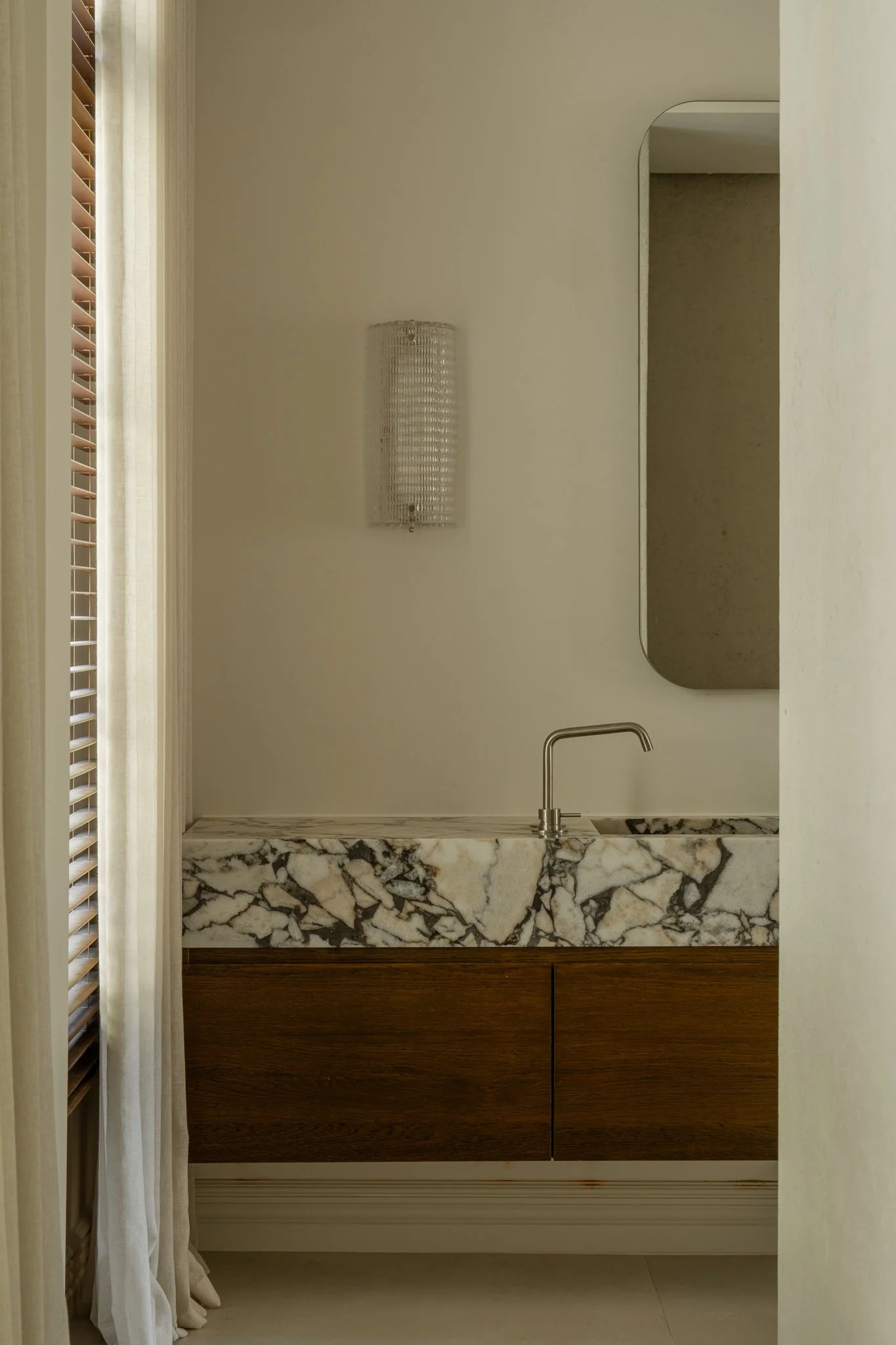 A bathroom vanity with a marble countertop, wooden cabinet, a modern faucet, a mirror, and a wall-mounted light fixture.