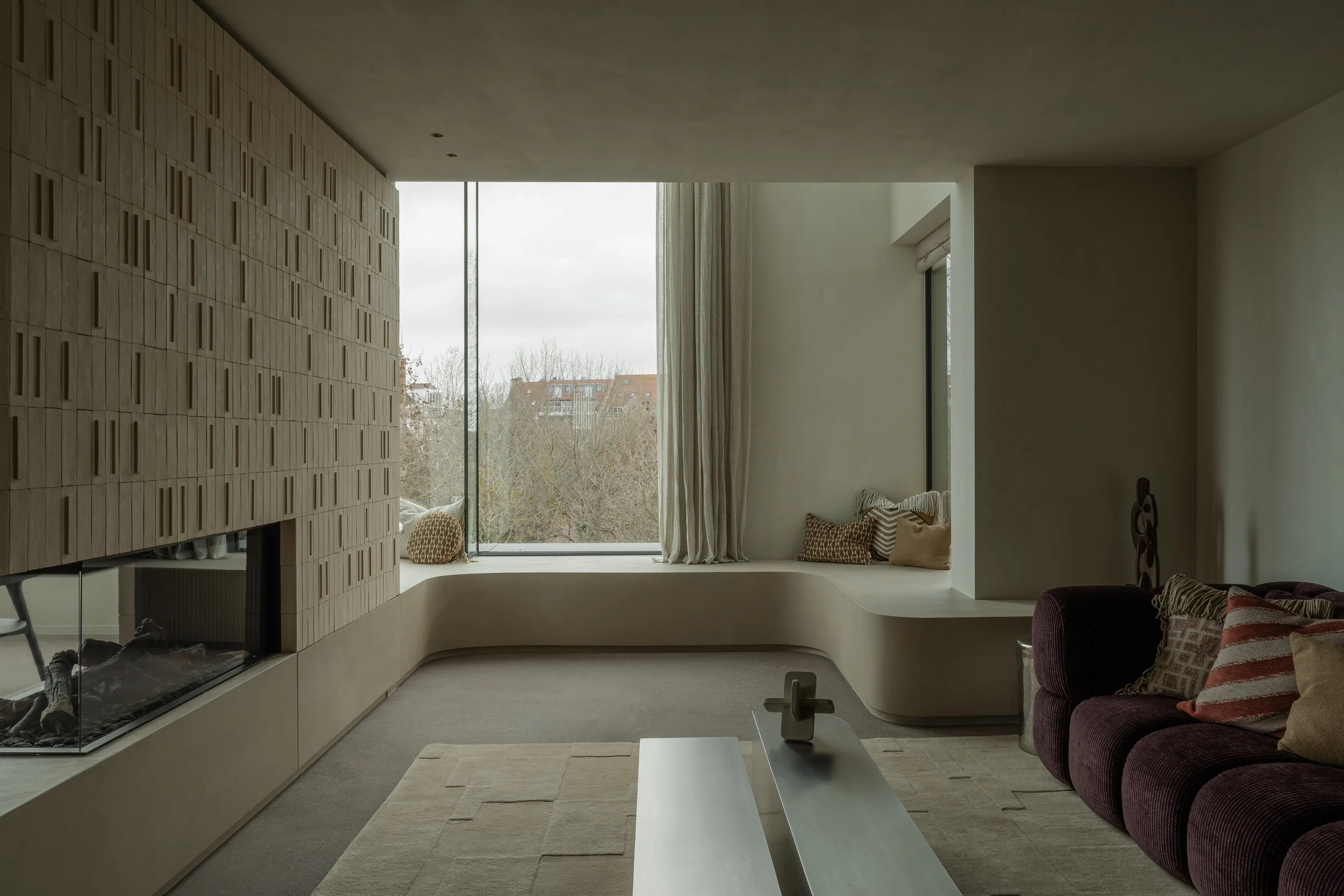 Living room with large window, beige bench seating area, purple sofa with patterned pillows, fireplace, and modern decor.