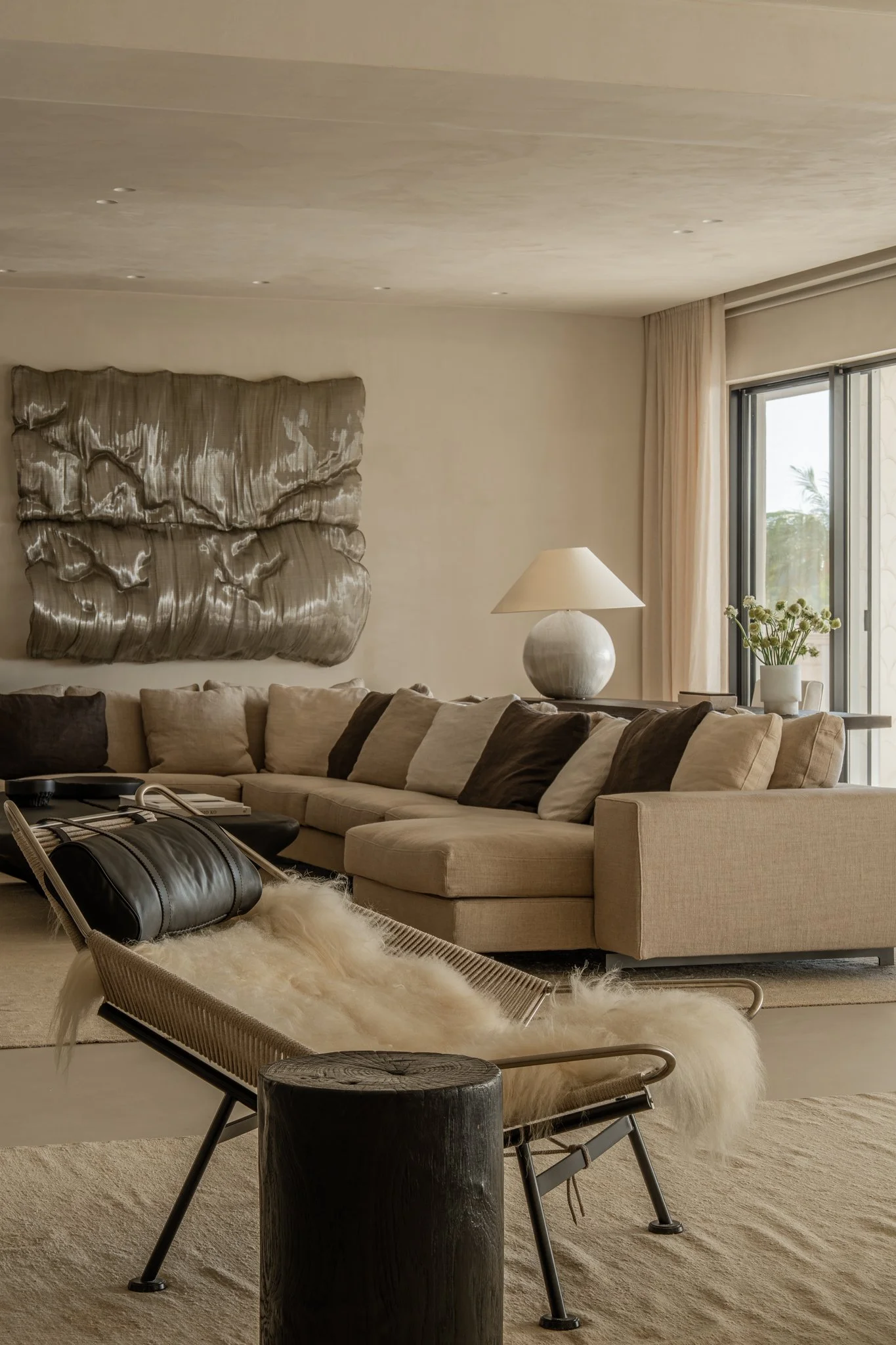 Living room with beige couch, dark brown and beige pillows, a leather bag on a chair with a sheepskin throw, a modern wall art piece, a table lamp on a side table, and a window with drapes.