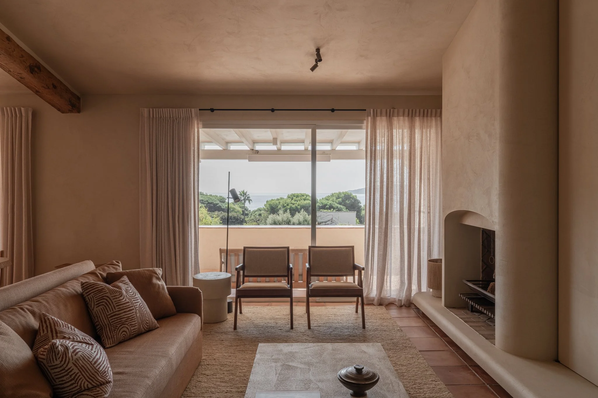 Living room with beige sofa, patterned pillows, two chairs facing a balcony, large sliding glass doors with sheer curtains, fireplace, and outdoor view of trees and sky.