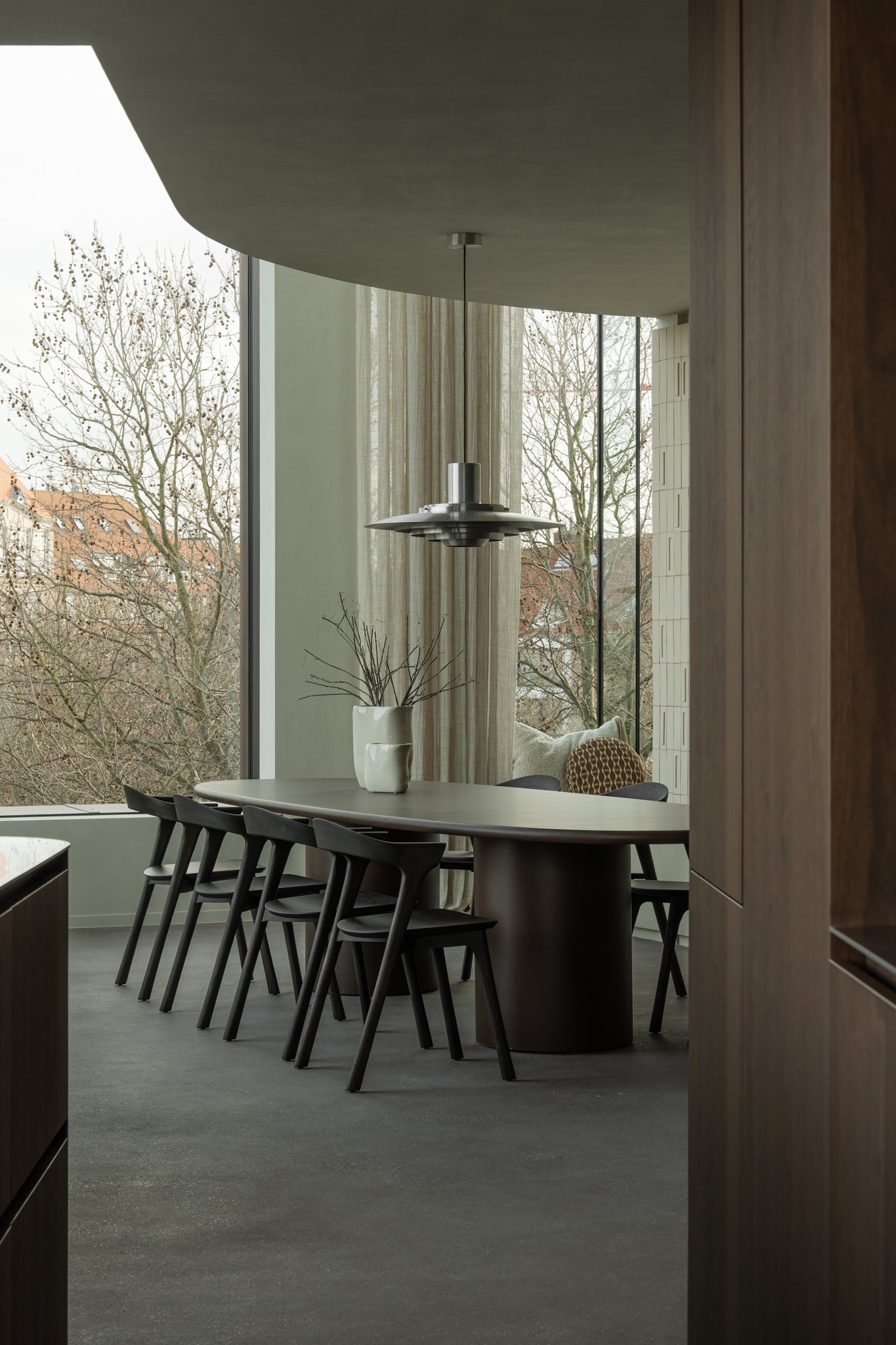 Modern dining room with a round table, six black chairs, a white vase with branches, large windows with cityscape views, textured curtains, and a sleek metallic light fixture.