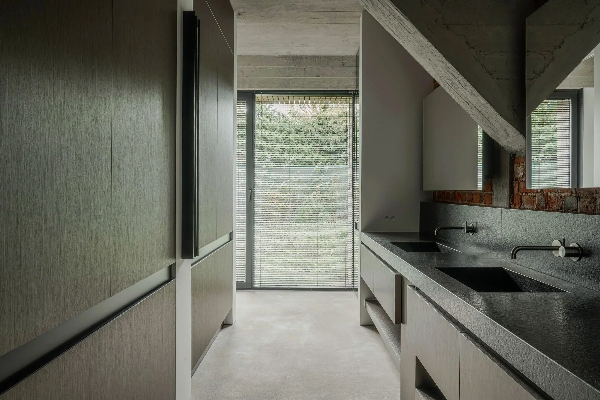 Modern kitchen with black countertops, gray cabinetry, and a large window with blinds showing trees outside.