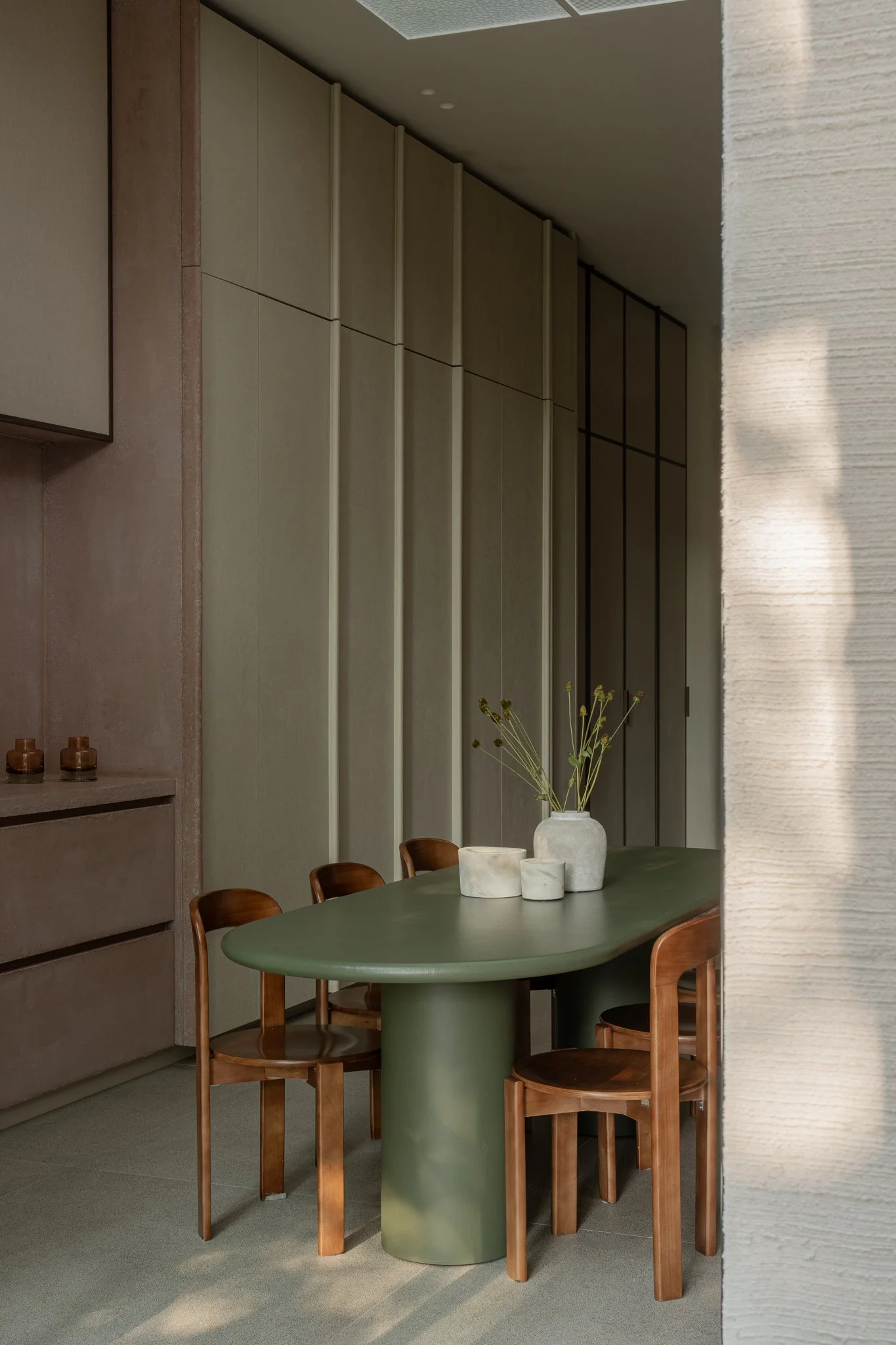 A dining area with four wooden chairs around an oval green table. A white vase with tall, thin flowers sits on the table, along with three smaller white vases. The background features a textured wall and built-in cabinetry.